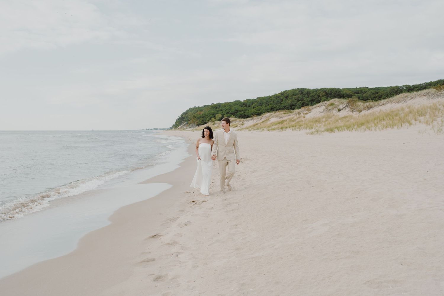 Couple walking together on a serene beach with sand dunes and ocean views in soft muted tones.