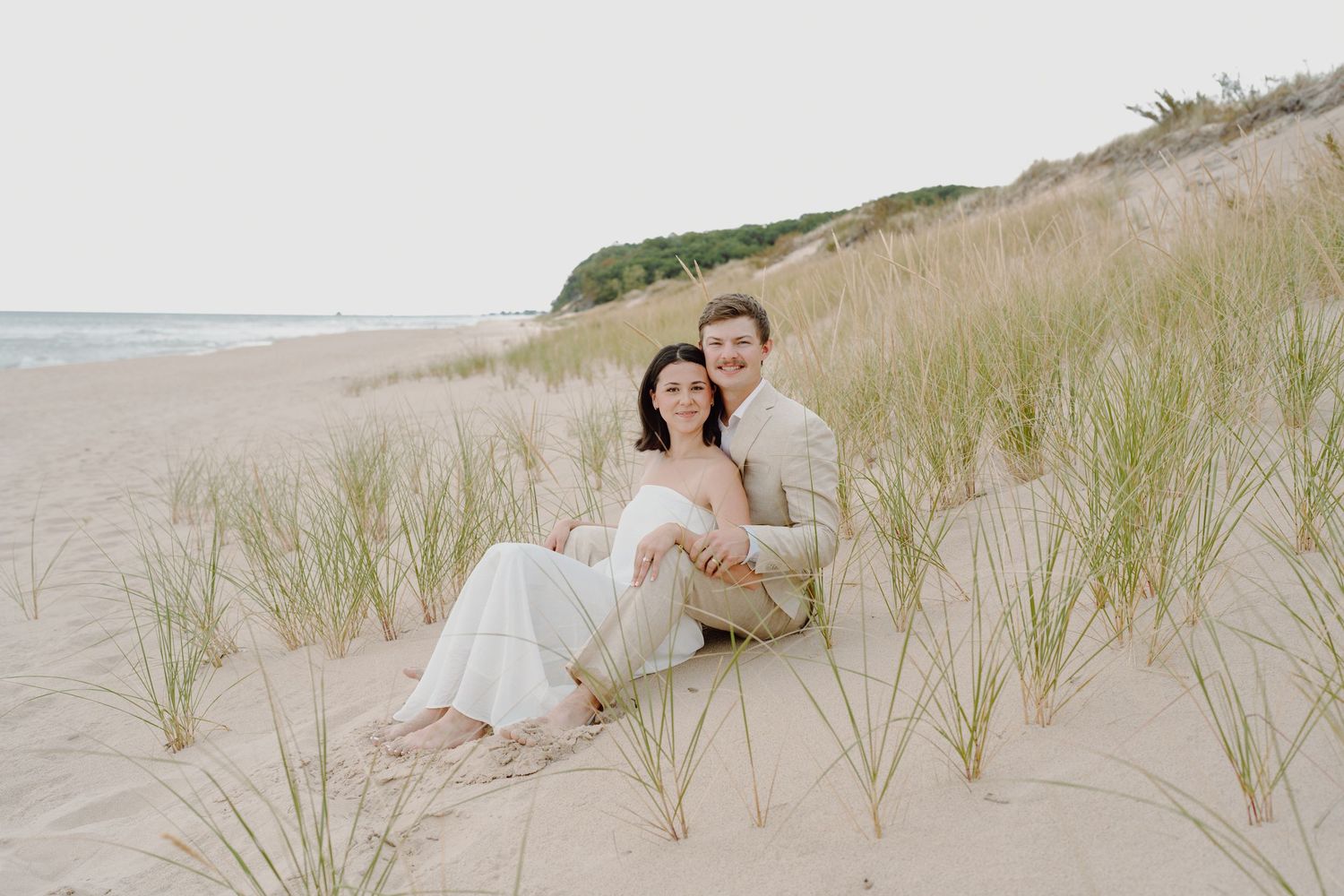Couple in white and beige lounging on sandy dunes with beach grass.