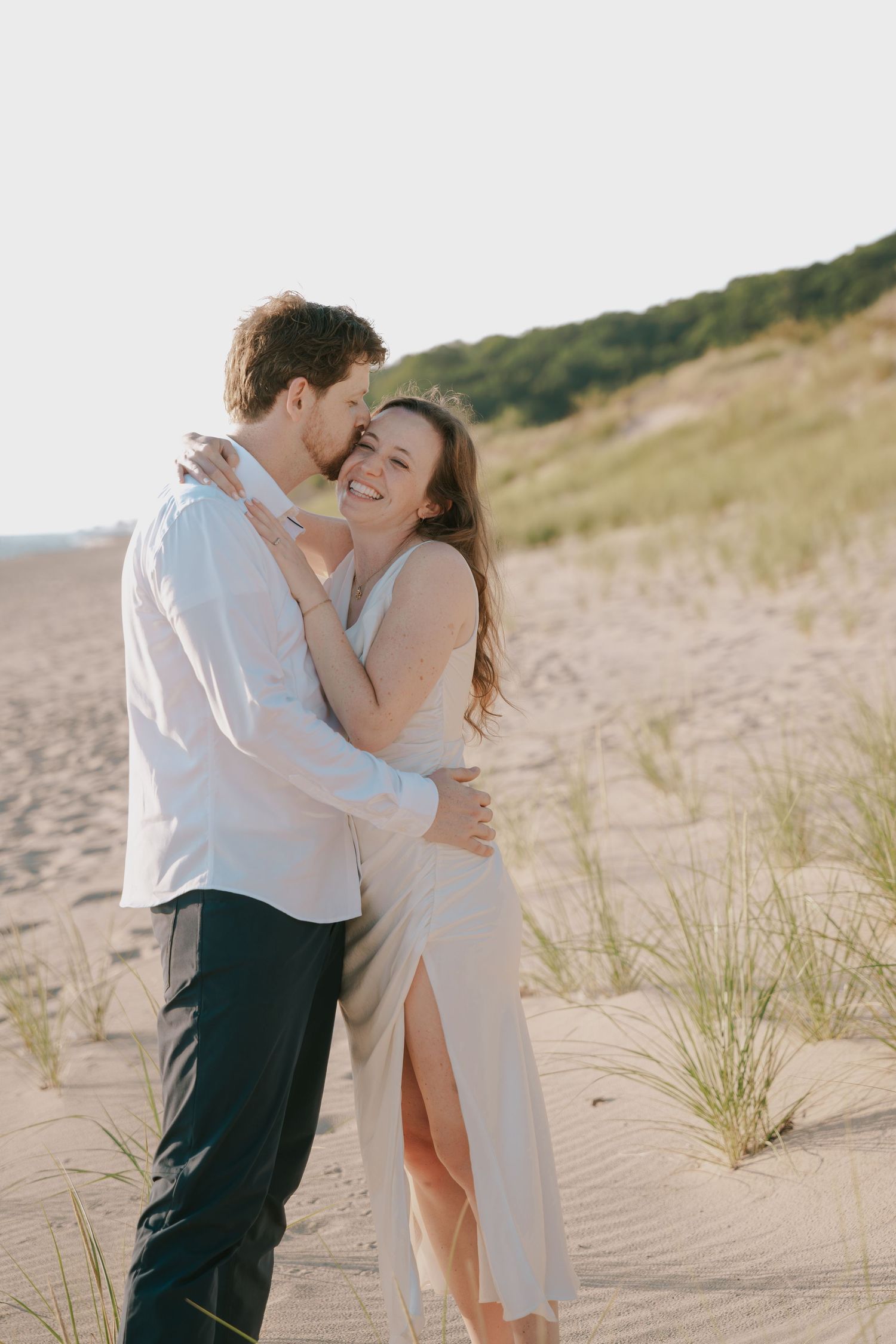 A tender moment shared between a couple on a beach with coastal dunes.