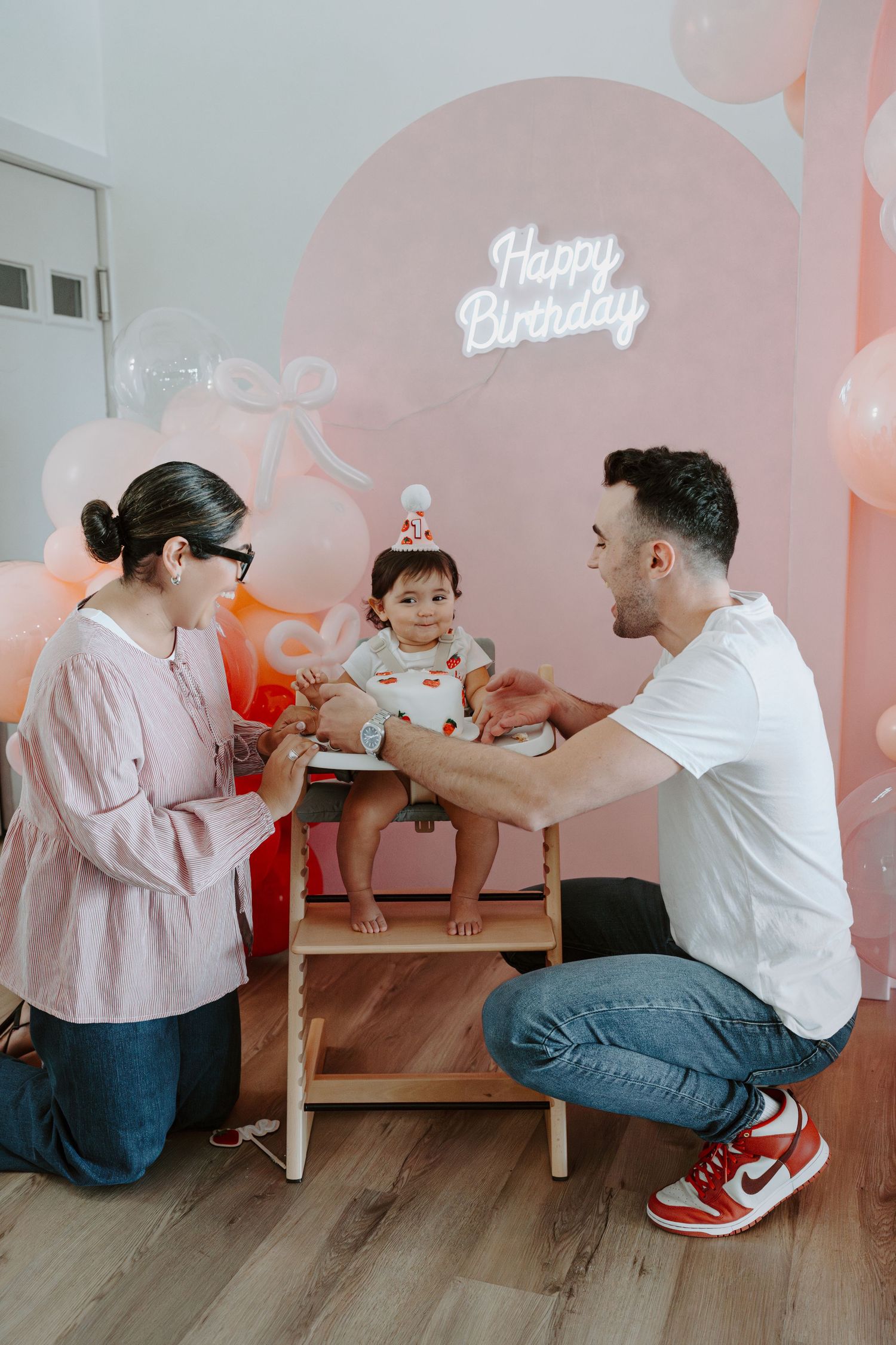 Birthday party scene with family sharing cake against pink balloon backdrop and neon signage.