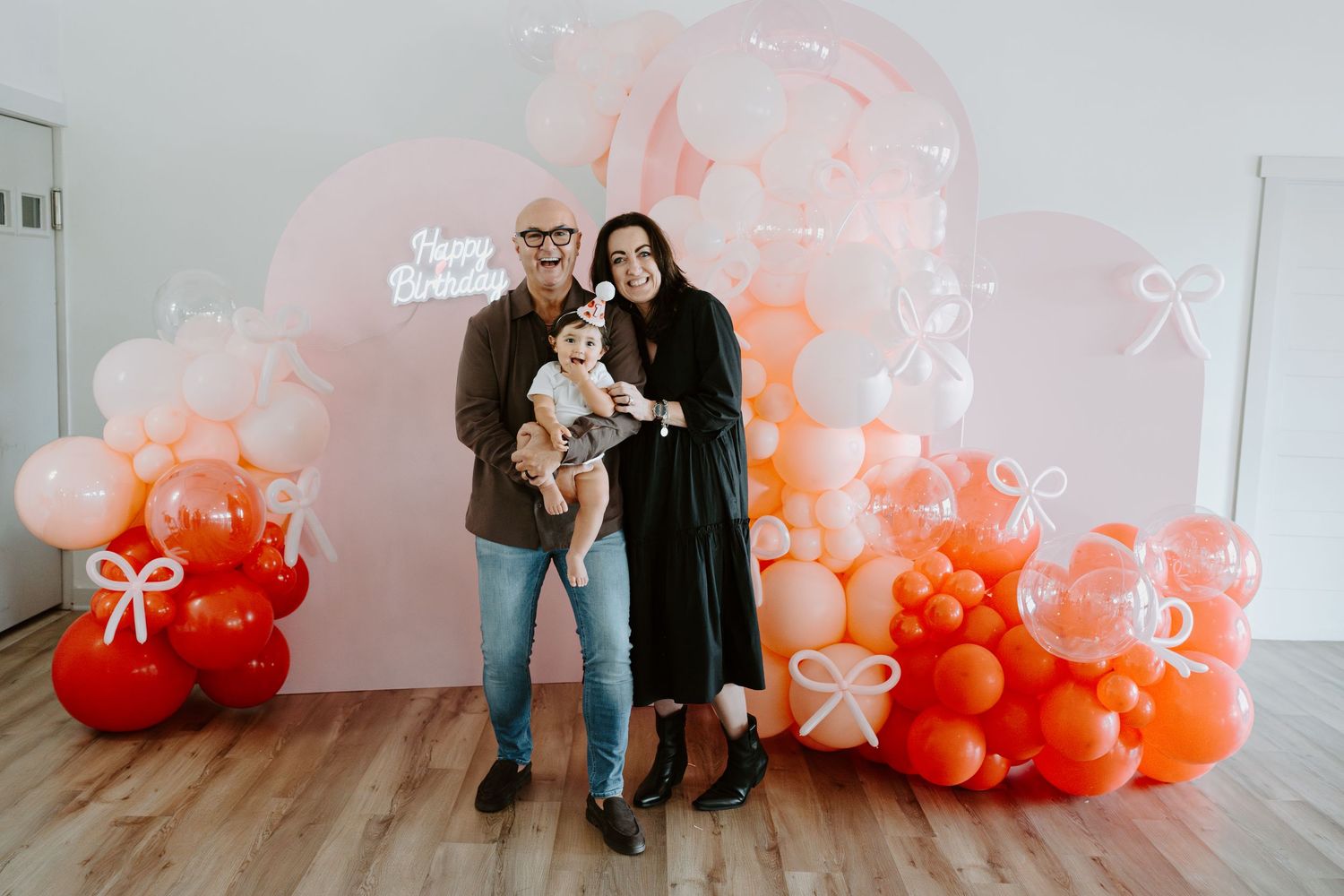 A couple holds their baby while standing before a pink and orange balloon arch.