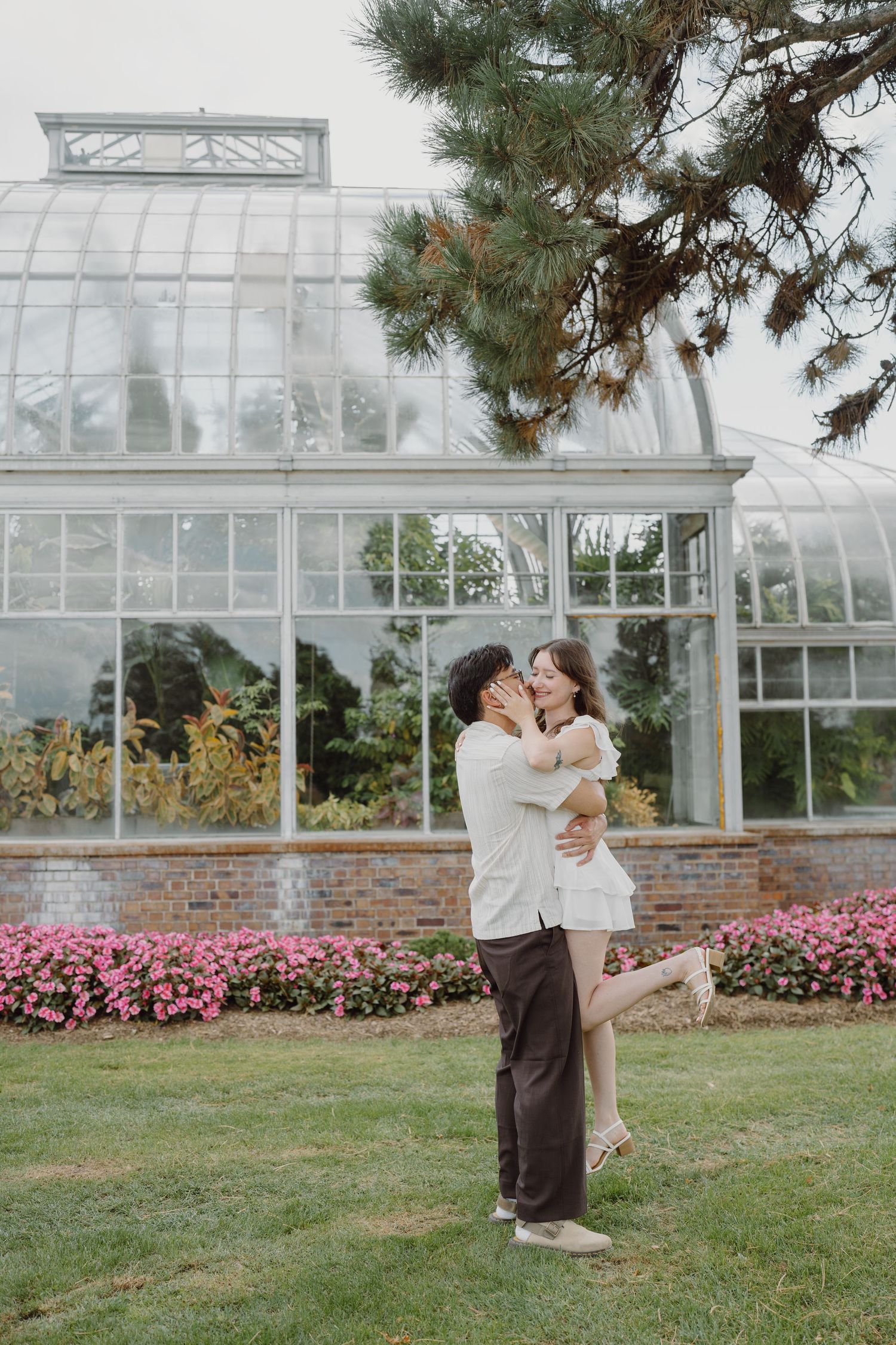 A tender moment captured between two people on the lawn with a greenhouse backdrop.