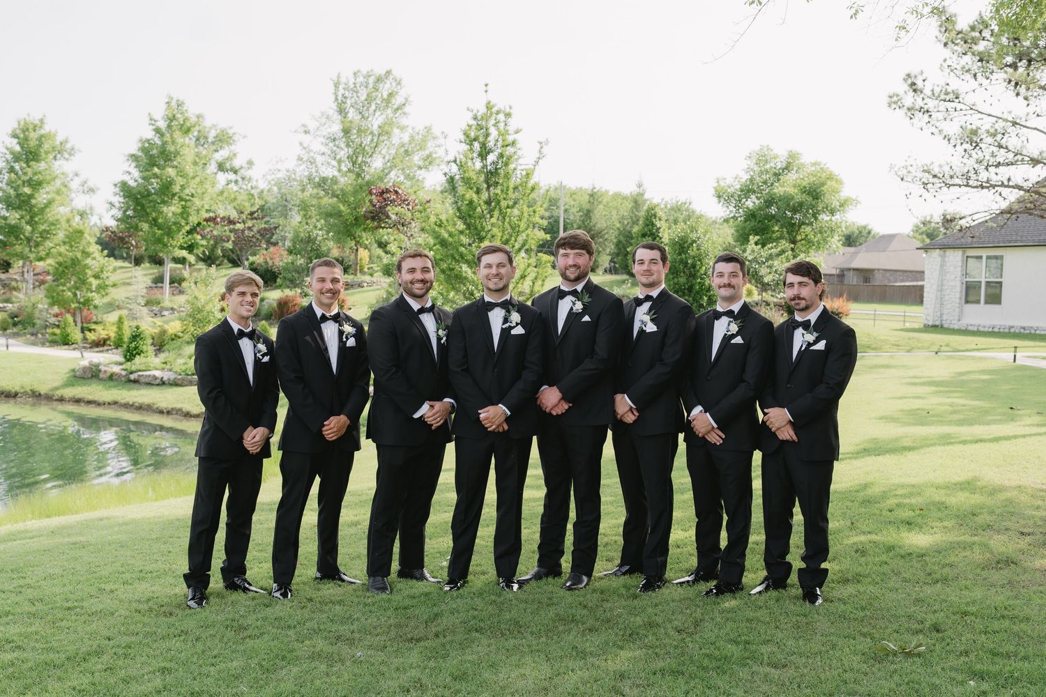 Groom and groomsmen in matching black tuxedos with white boutonnieres posing outdoors on green lawn at wedding.