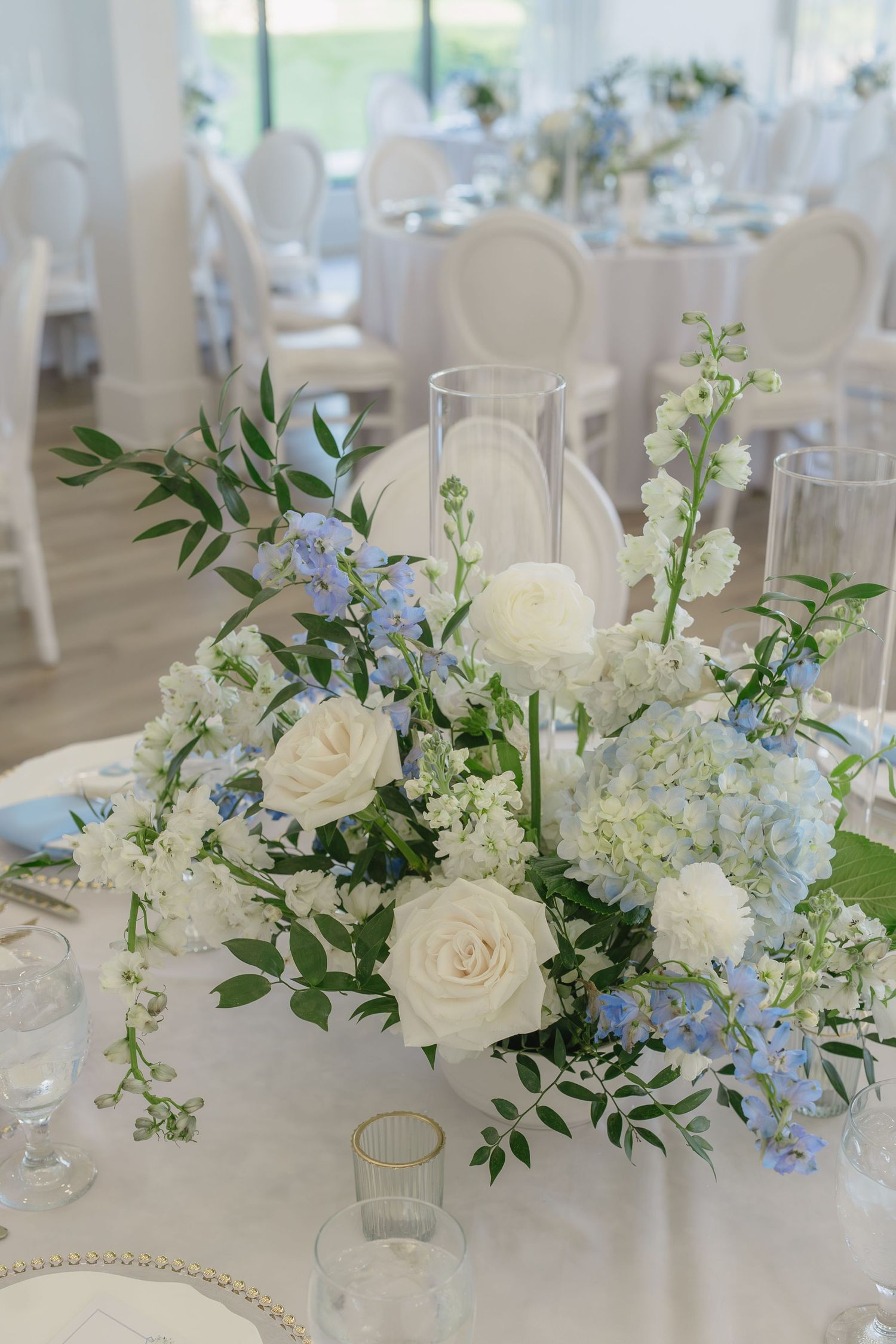Elegant wedding reception centerpiece with white roses, blue hydrangeas, and greenery on a white tablecloth.
