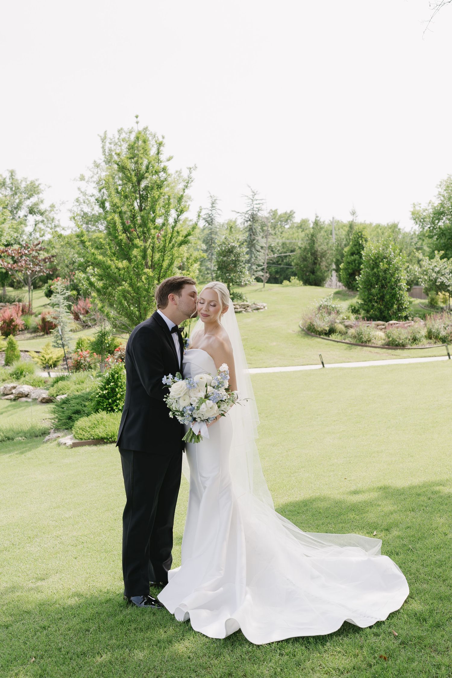 A bride and groom share a romantic moment on a lush green lawn, with the bride holding a white bouquet.