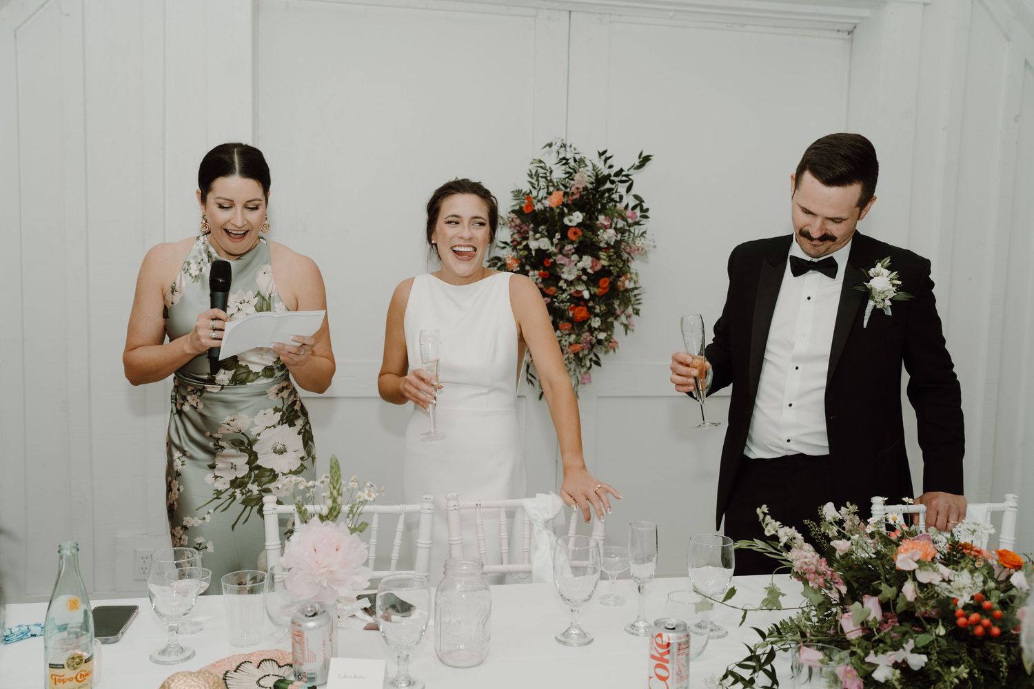 Wedding party celebrates with laughter and drinks in an elegant white reception room.