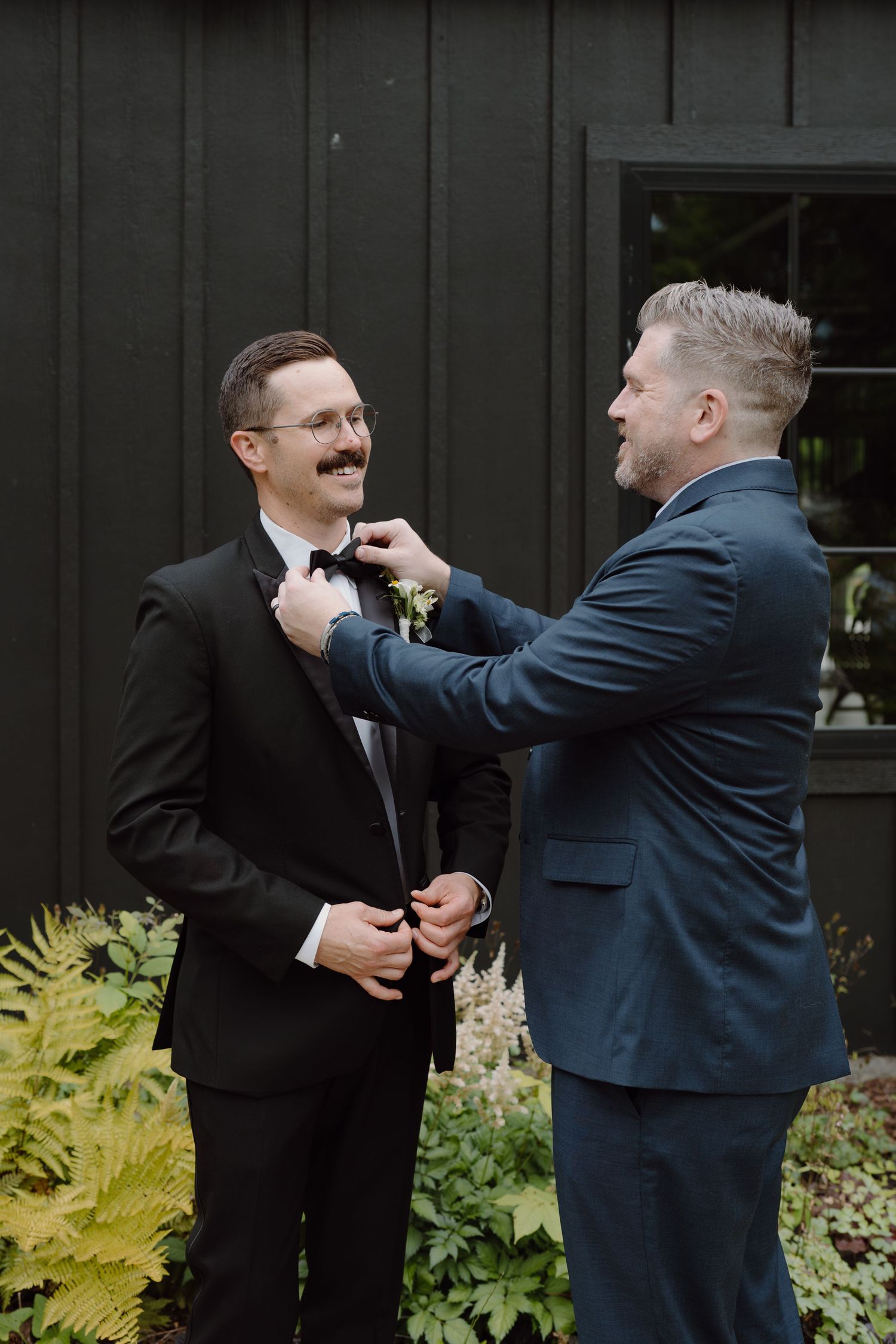 Groomsmen assist each other with boutonniere pinning in an outdoor setting.