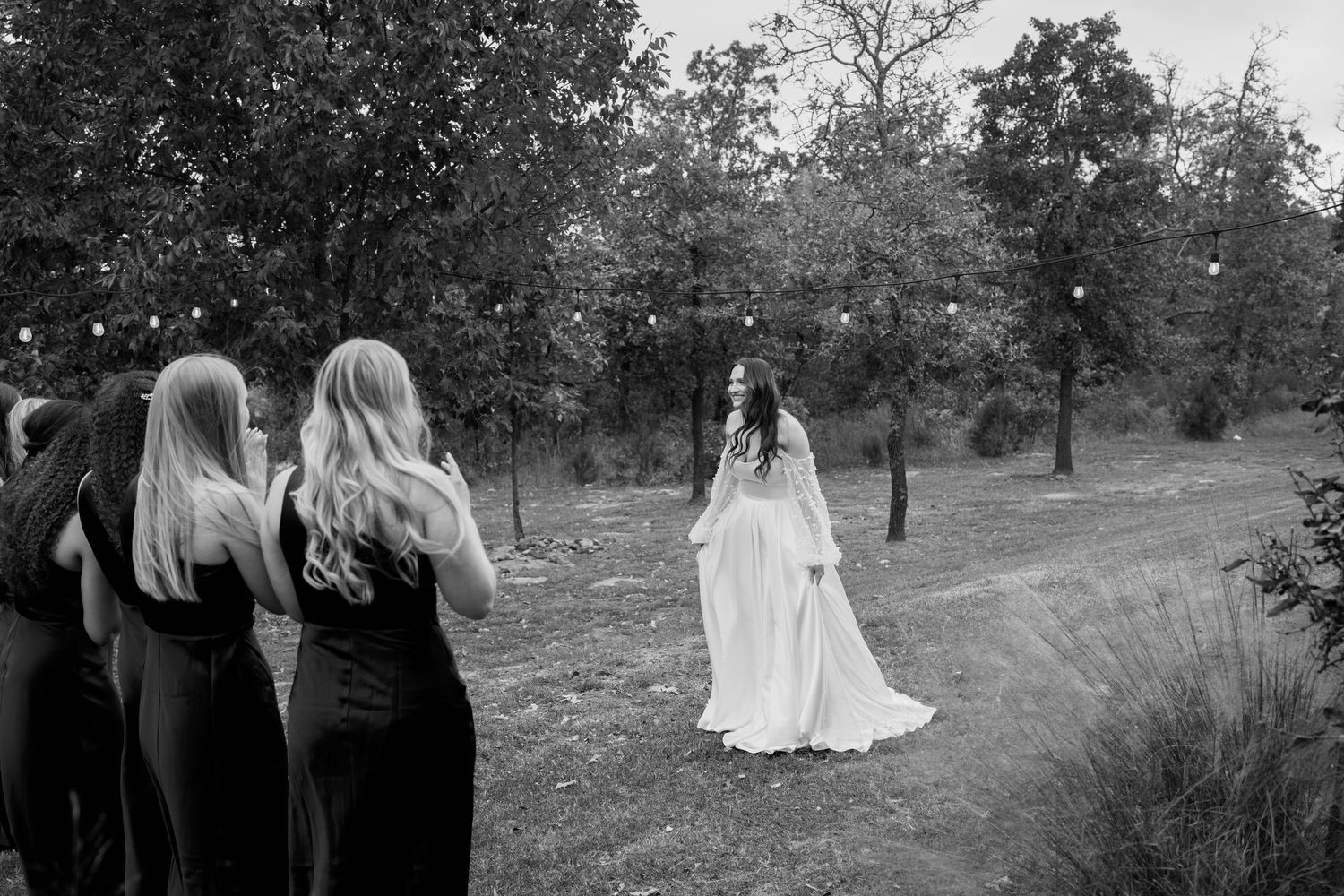 Black and white photo of bridal party watching someone pose in a flowing white dress outdoors.