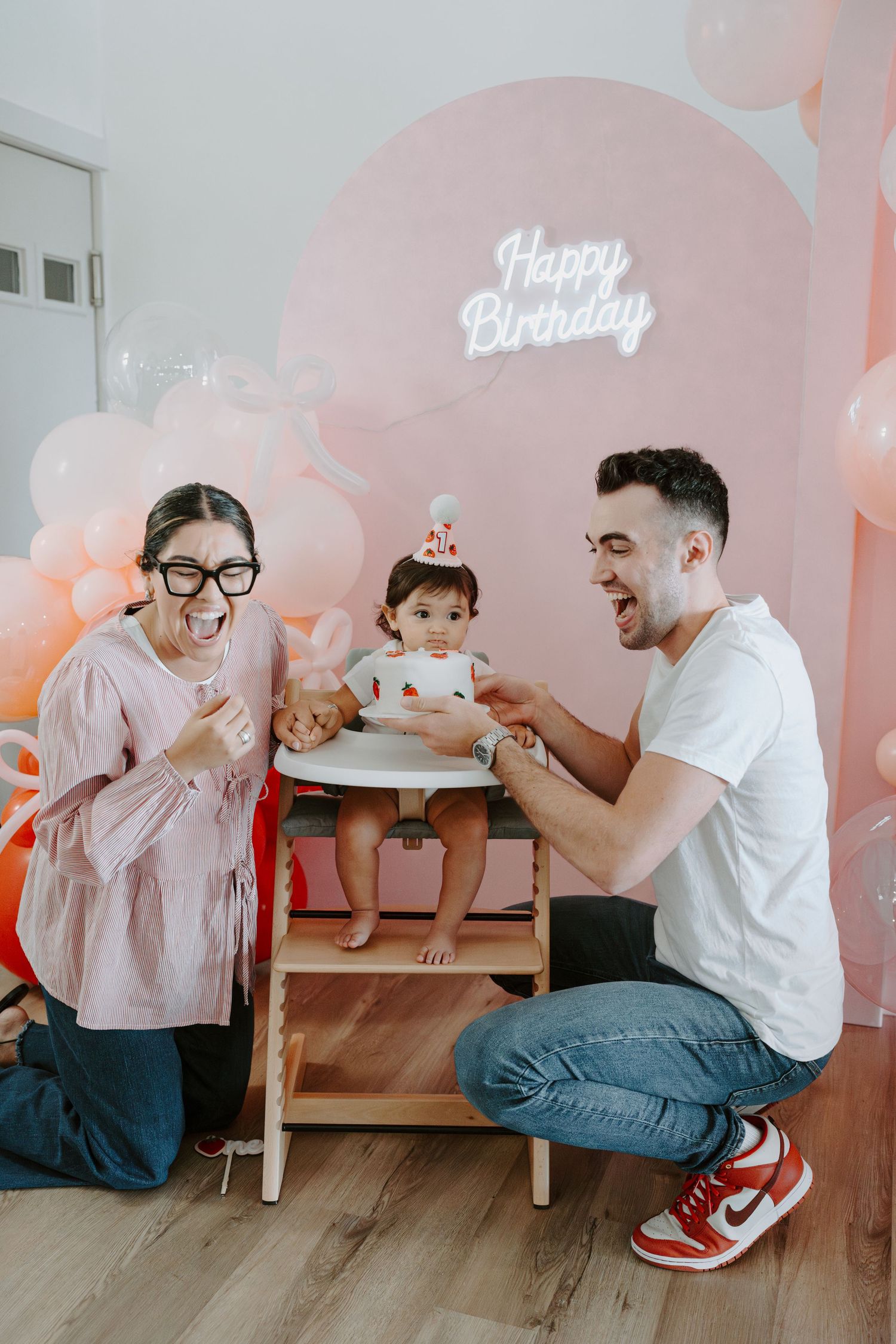 Family celebrating a birthday with cake on wooden table against pink balloon backdrop and neon sign.