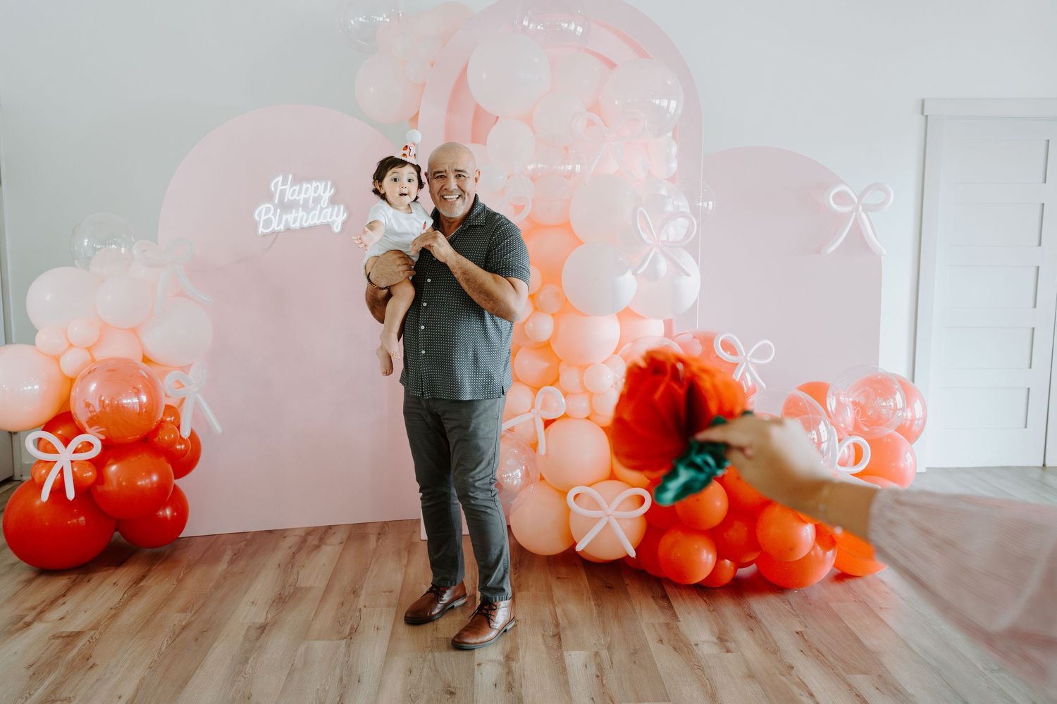 Birthday party setup with pink and coral balloon arch against white wall and wooden floor.