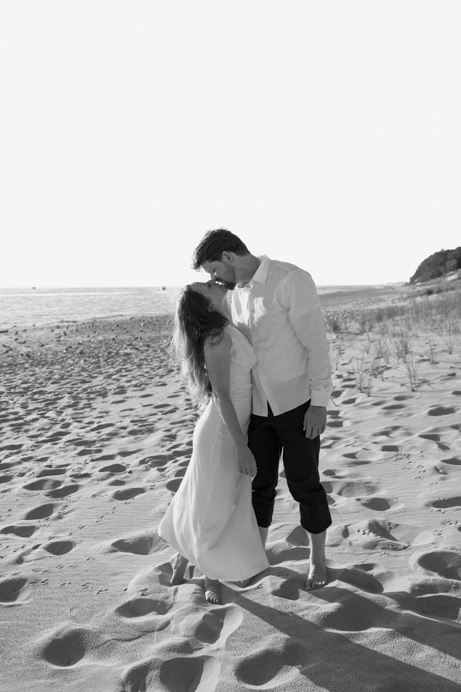 A romantic black and white photo of a couple embracing on a sandy beach with dunes in the background.