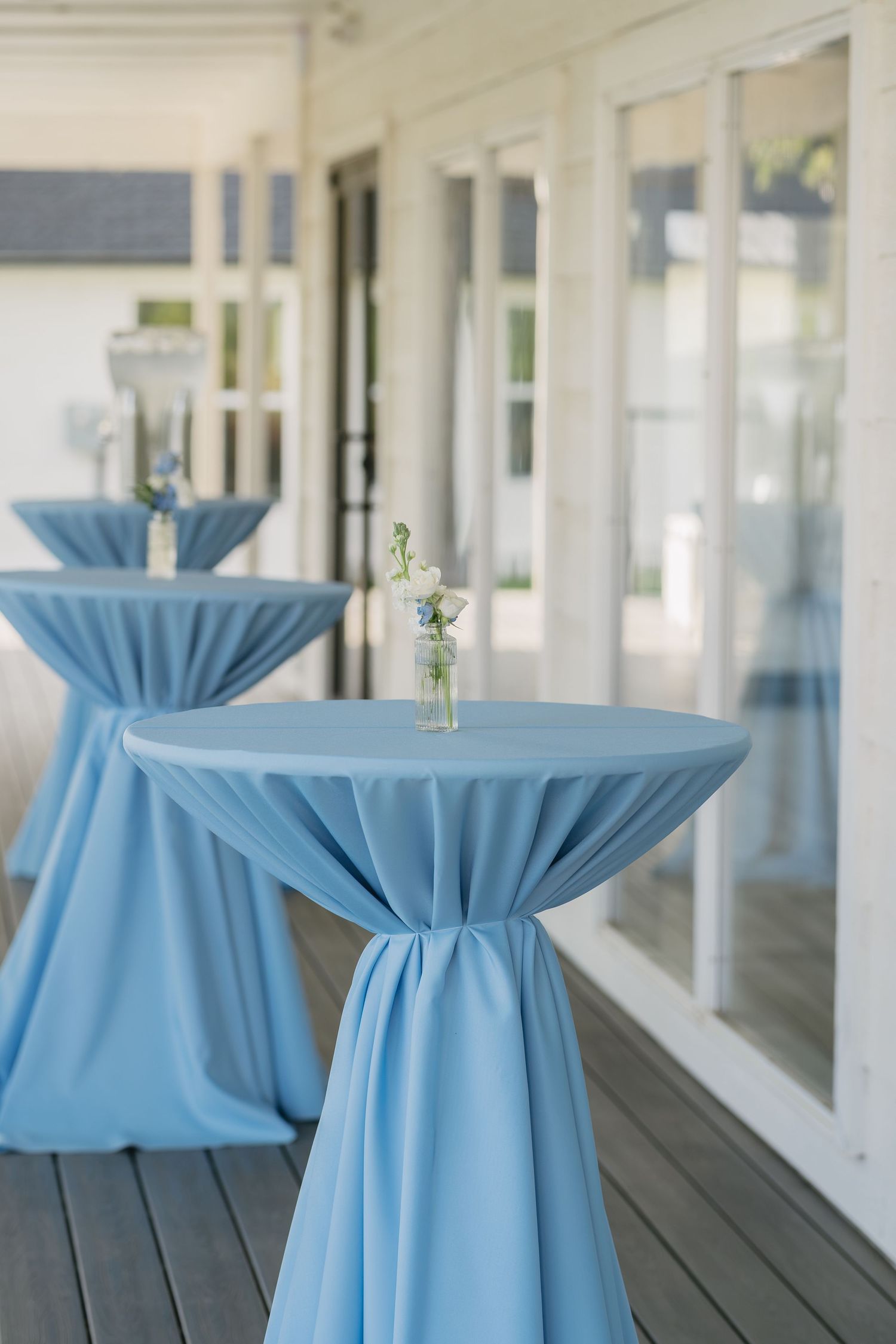 Elegant cocktail tables draped in light blue fabric with small floral centerpieces on an outdoor patio.