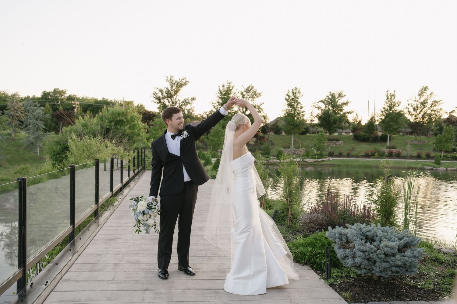 Bride and groom dancing on a scenic garden bridge near a pond, bride in white gown, groom in black tuxedo.