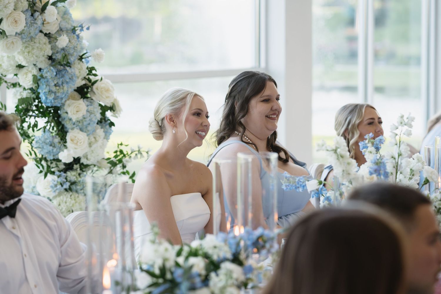 Bride and bridesmaid laughing at wedding reception with elegant blue and white floral arrangements on decorated table.