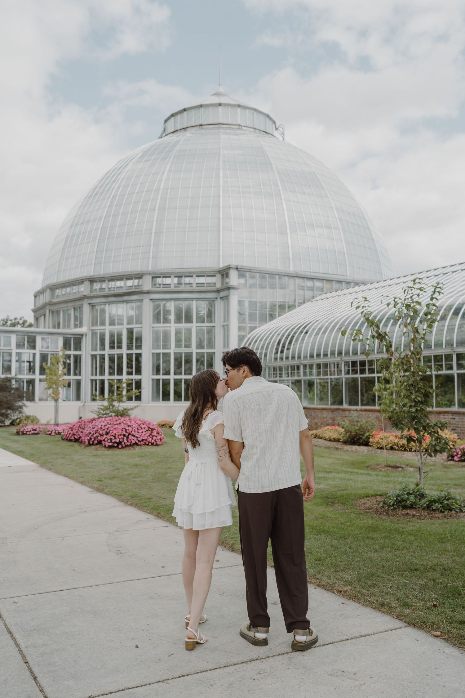 Two people walk together near a conservatory building with pink flowers blooming nearby.