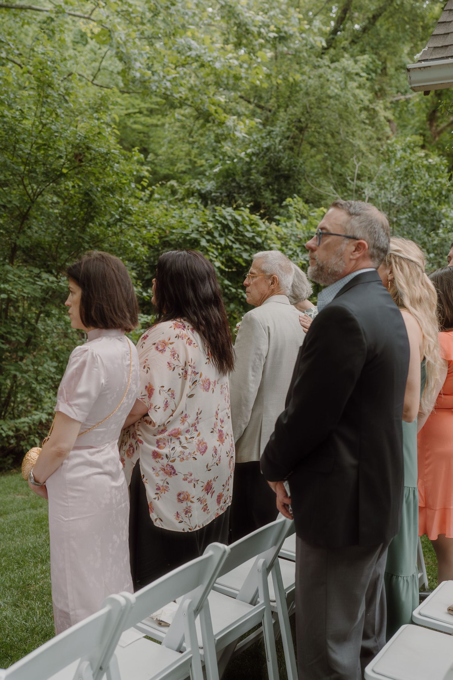 Wedding guests stand during an outdoor ceremony surrounded by lush greenery.