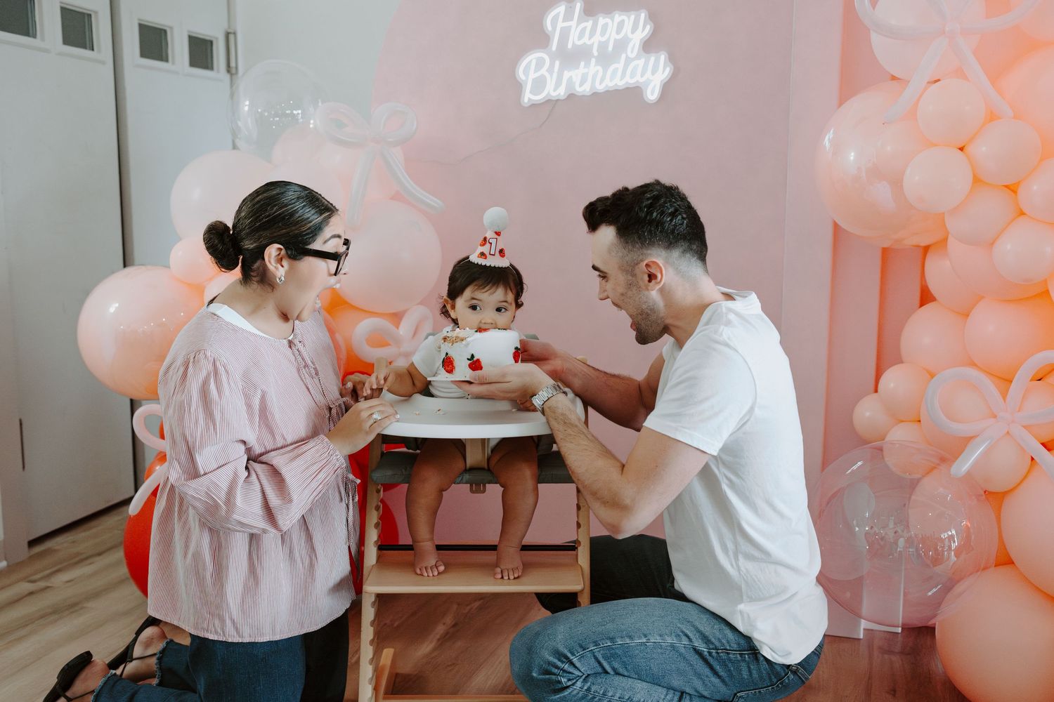 Birthday celebration scene with pink balloon wall and neon sign as backdrop for family gathering.