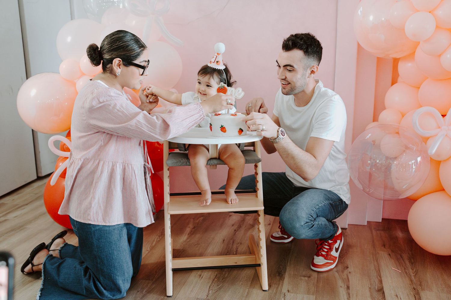 Parents kneeling beside baby in high chair during birthday celebration with pink balloon backdrop.