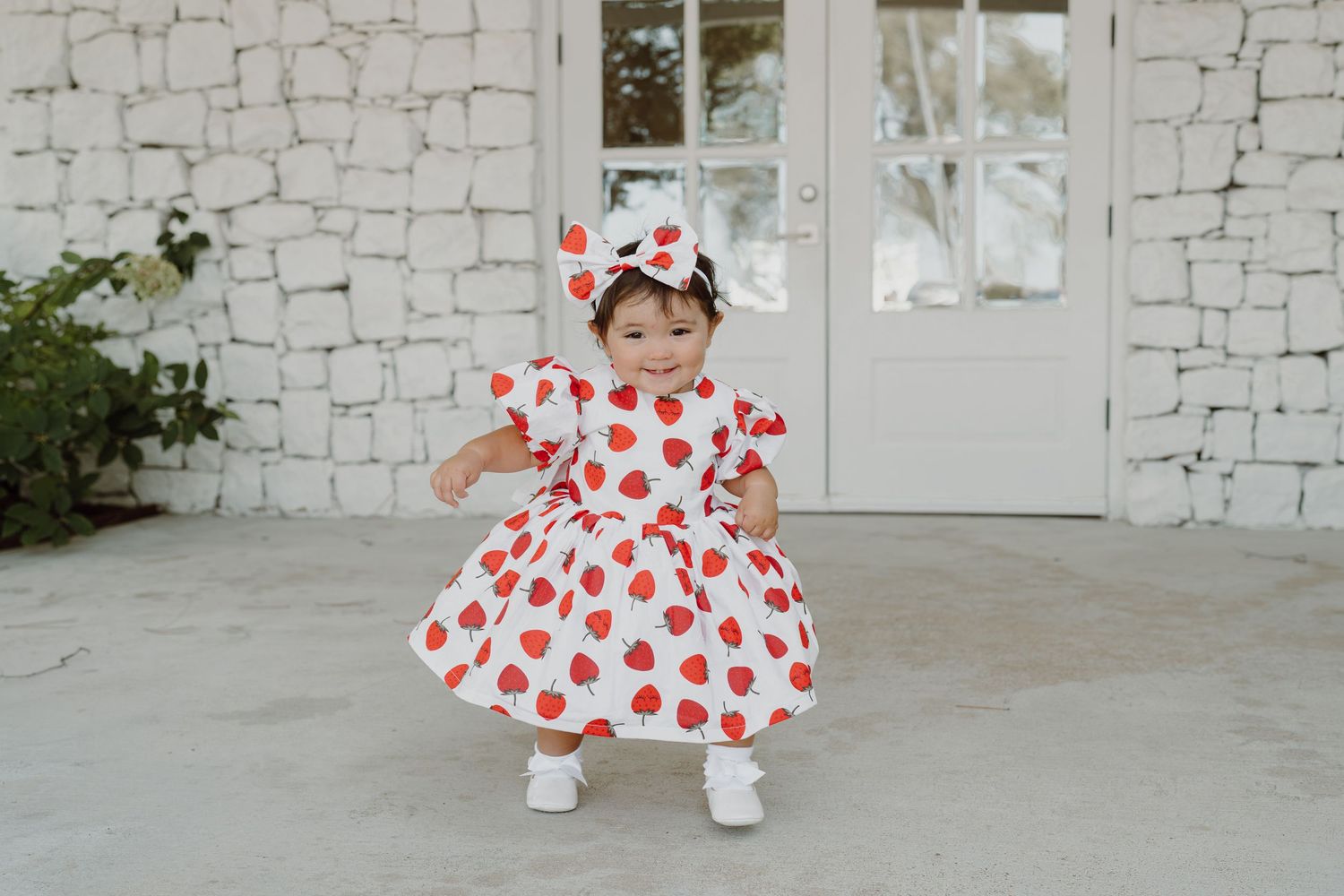 Playful pose in a white dress with red heart pattern against stone wall background.