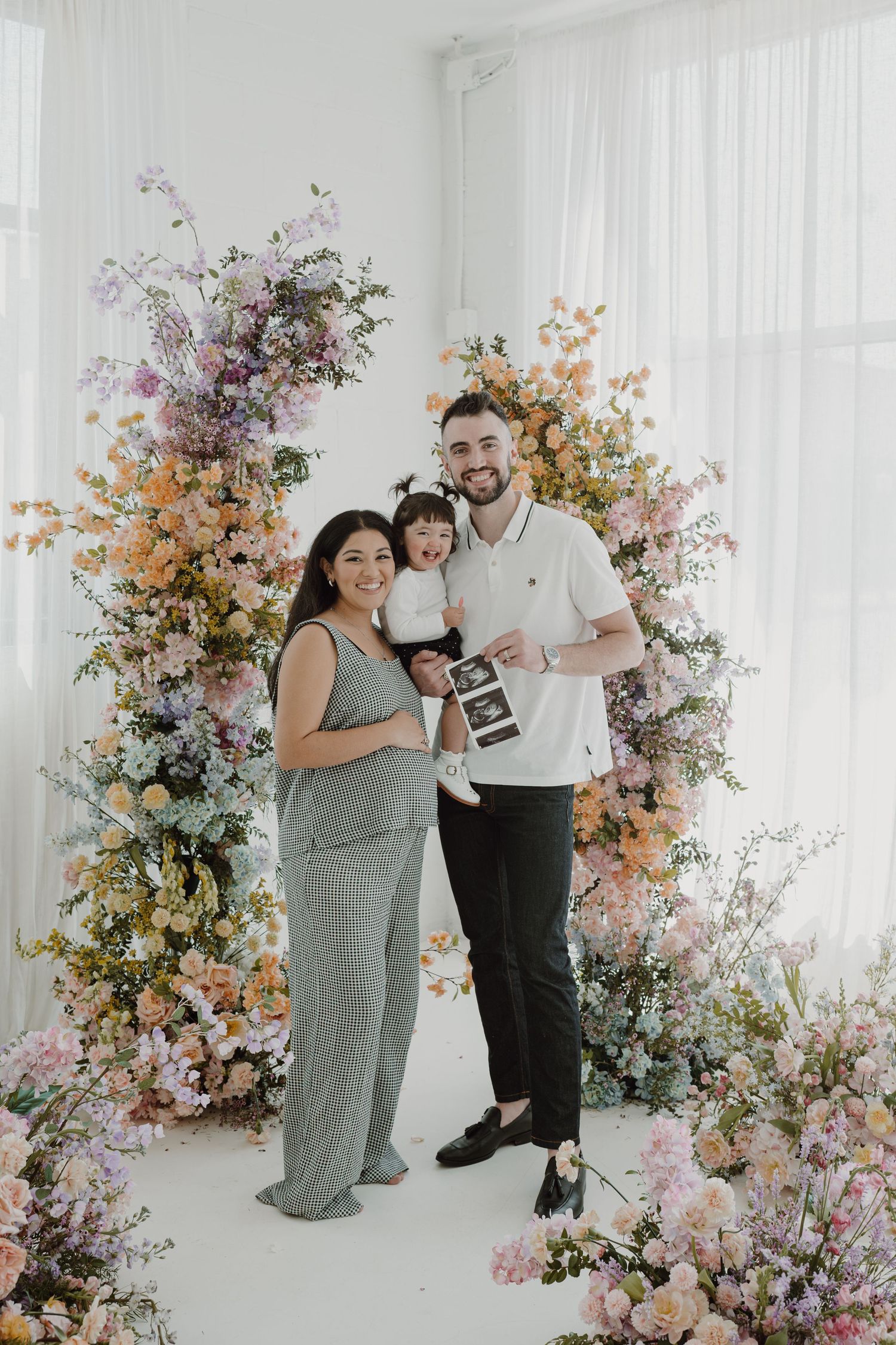 A growing family poses together against a pastel floral backdrop during a pregnancy announcement.