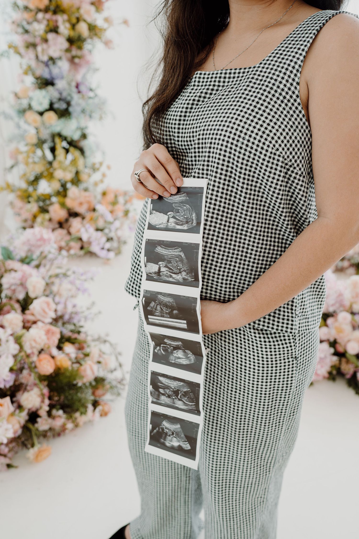 Pregnant woman in gingham jumpsuit holds ultrasound photos against floral backdrop.