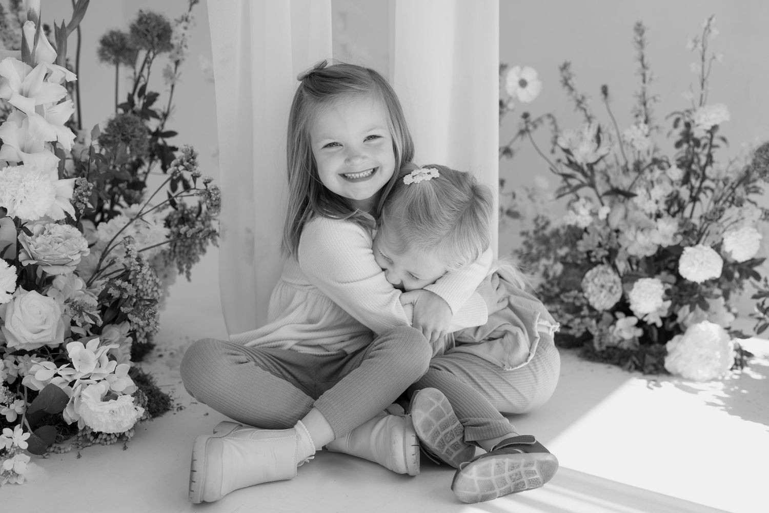 Black and white candid moment of siblings embracing amid floral decorations.