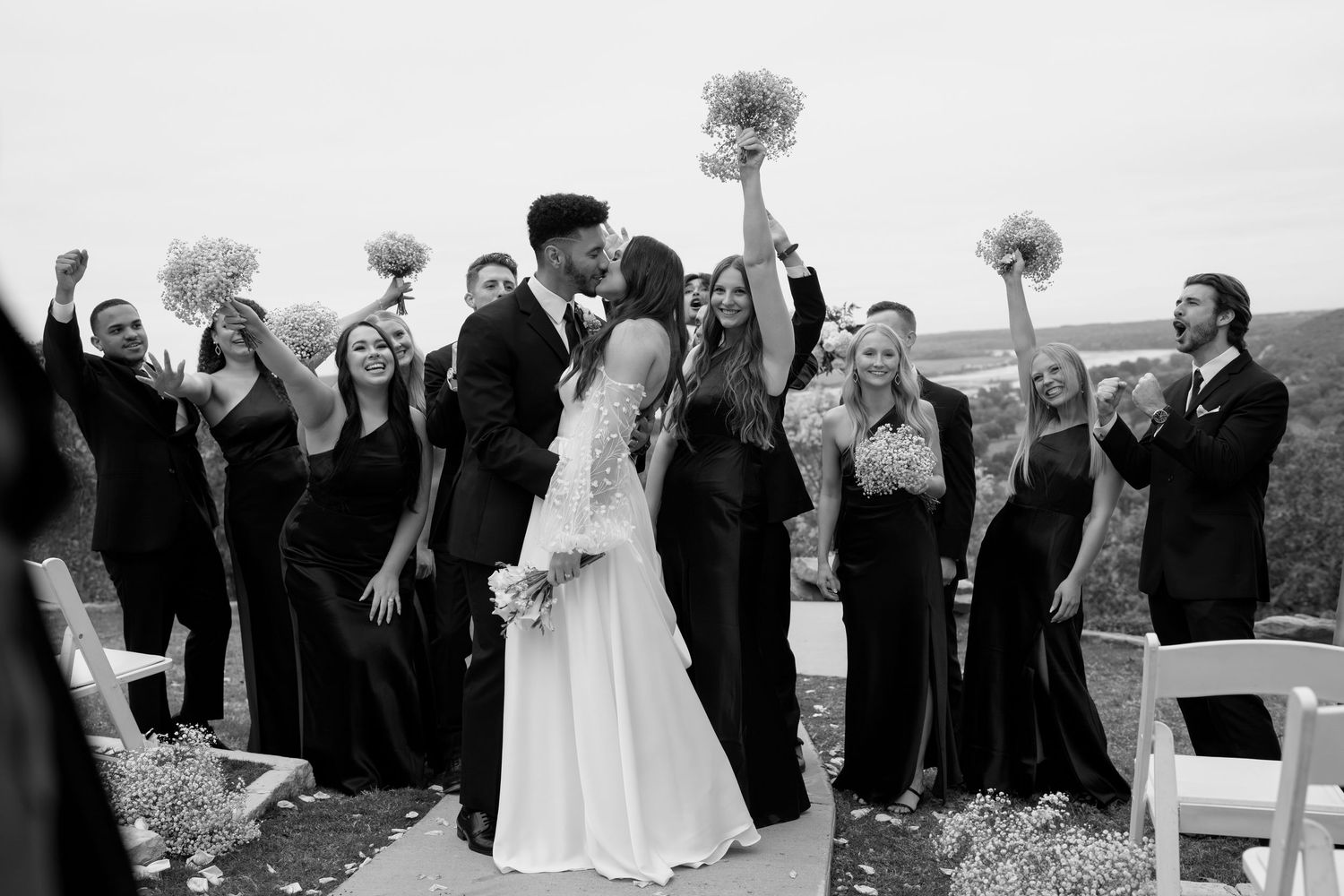 Wedding party celebrates with raised bouquets against a cloudy sky backdrop.