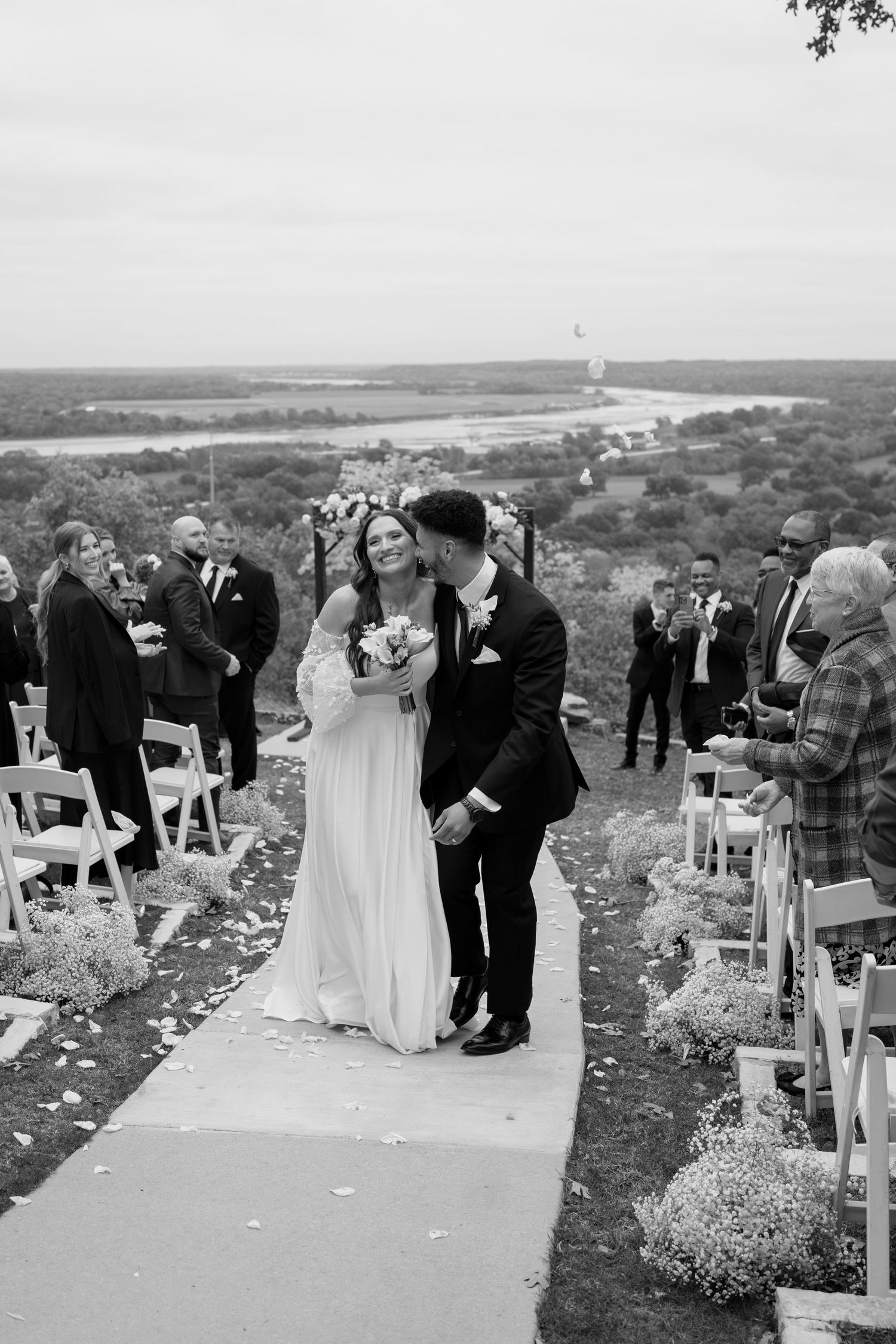 A black and white wedding ceremony photo overlooking a scenic valley with white chairs and flower petals.
