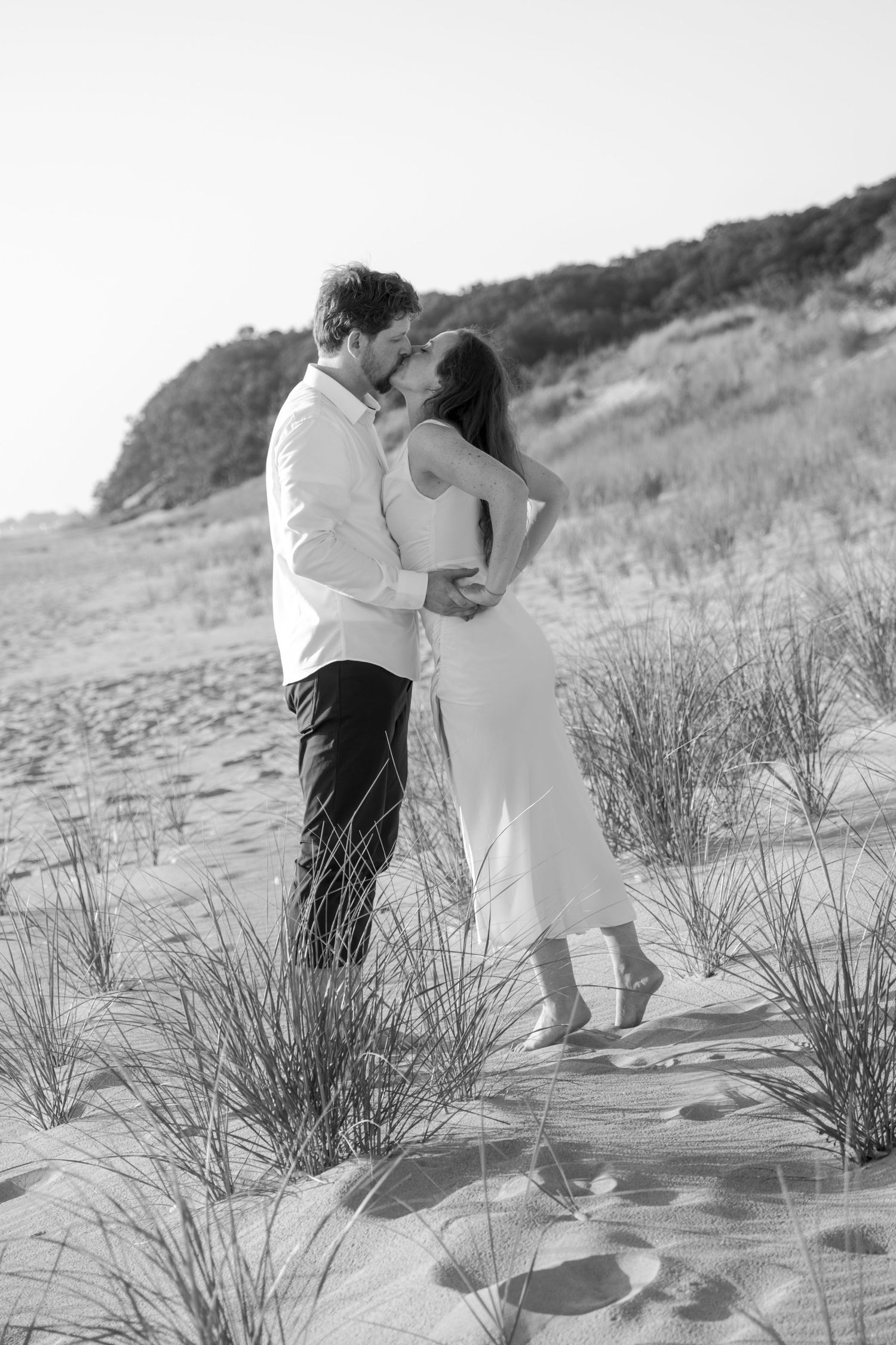 A tender moment between a couple in white clothing against a backdrop of beach dunes.
