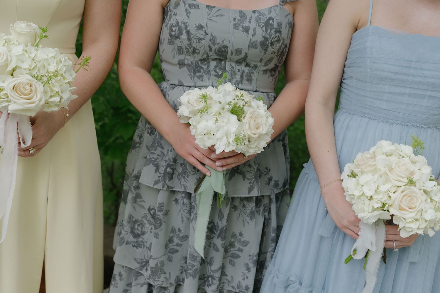 Three bridesmaids in blue and yellow gowns holding elegant white rose and hydrangea bouquets at a wedding ceremony.