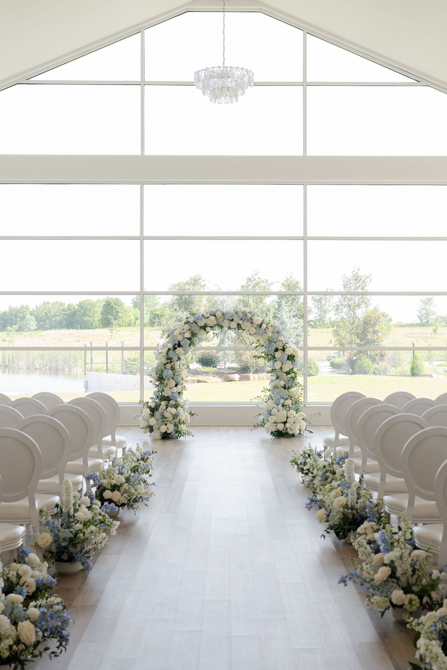 Elegant wedding chapel aisle with floral arch, blue and white flower arrangements, white pews, and large windows overlooking greenery.