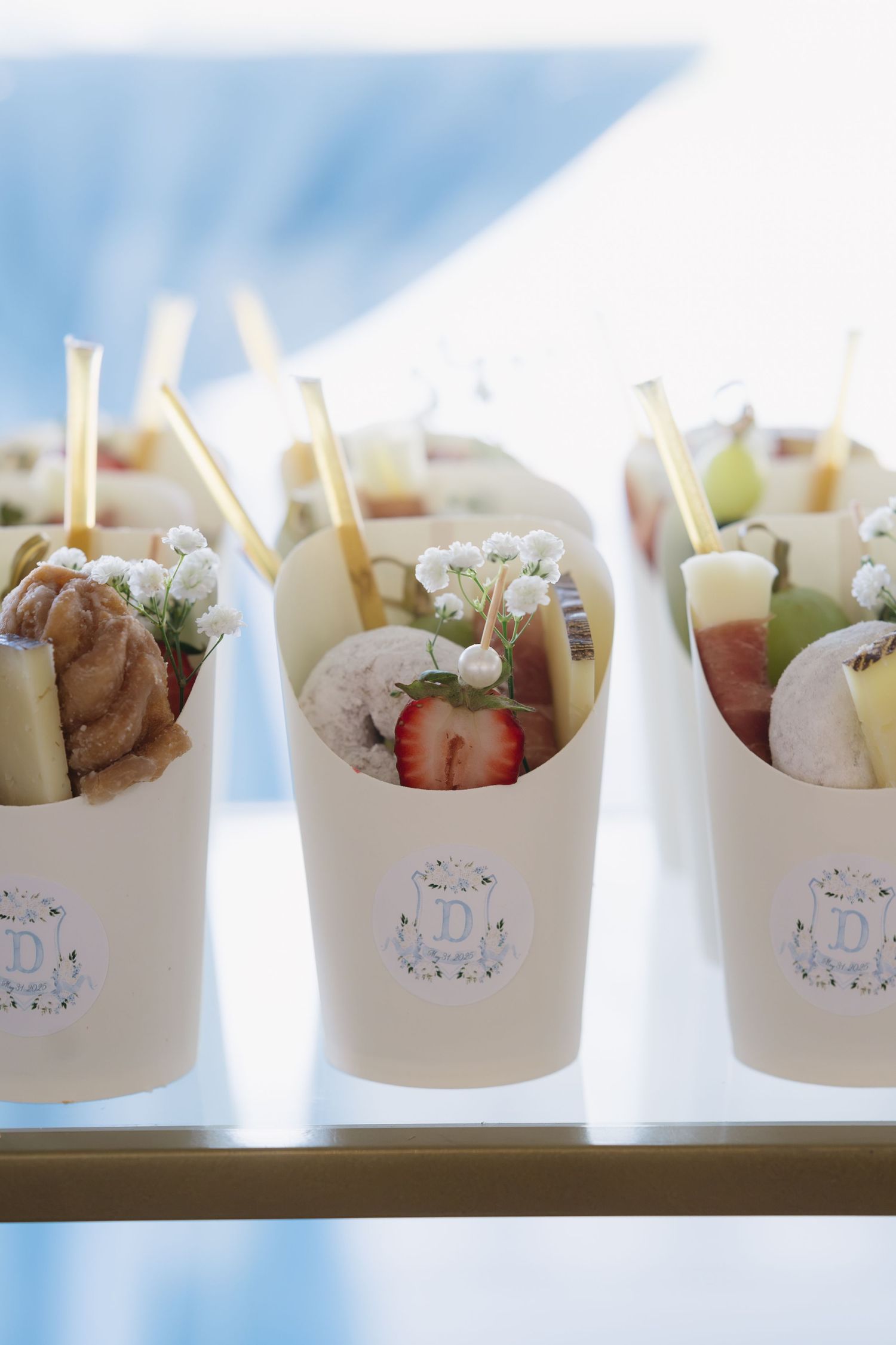Elegant white paper cups filled with fresh fruit, cheese, and gold straws displayed on a glass shelf.