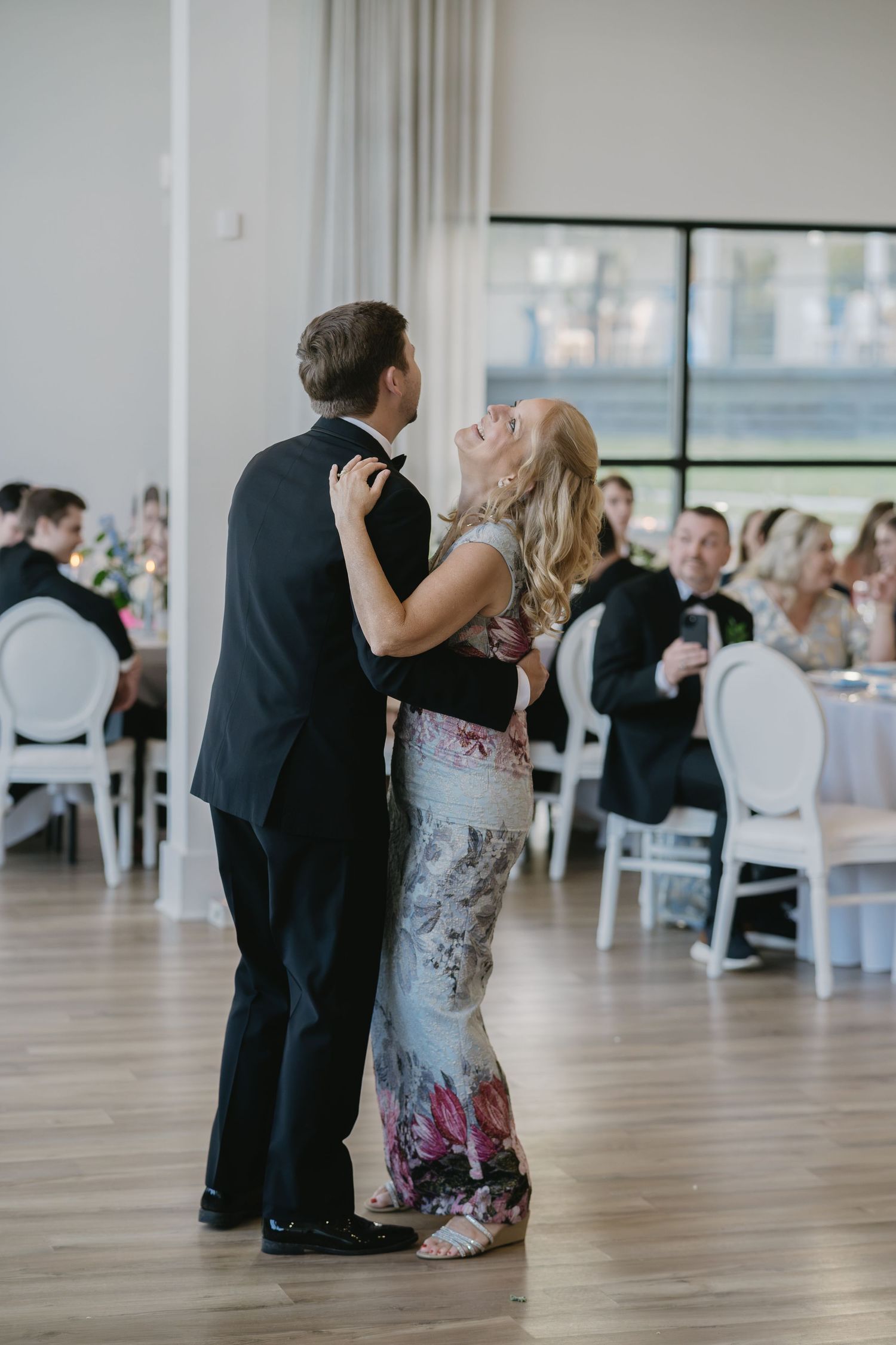 A couple shares a joyful mother-son dance at an elegant wedding reception in a bright, modern venue.