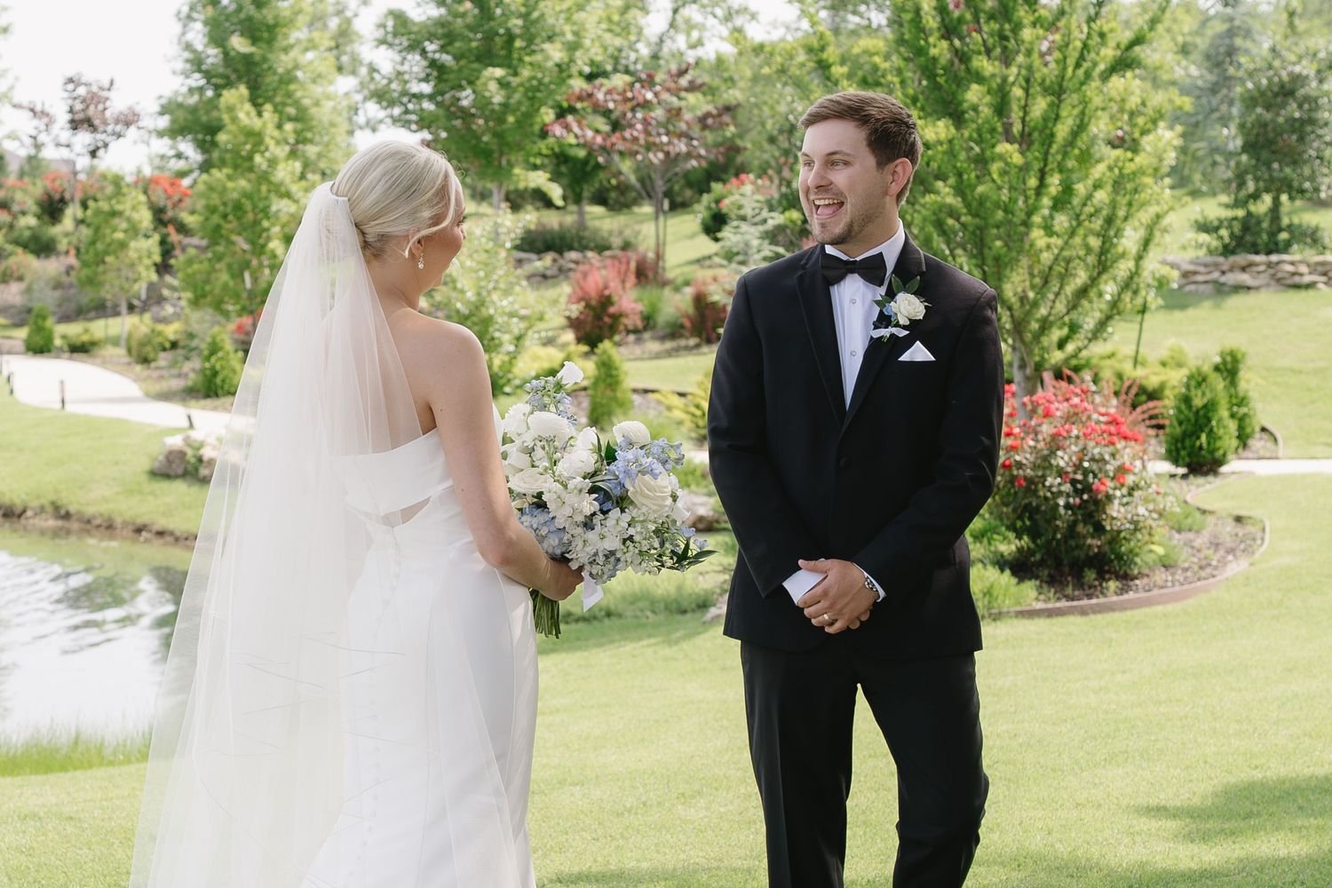 Bride in white gown and veil holds bouquet while groom in black tuxedo smiles at her outdoors.