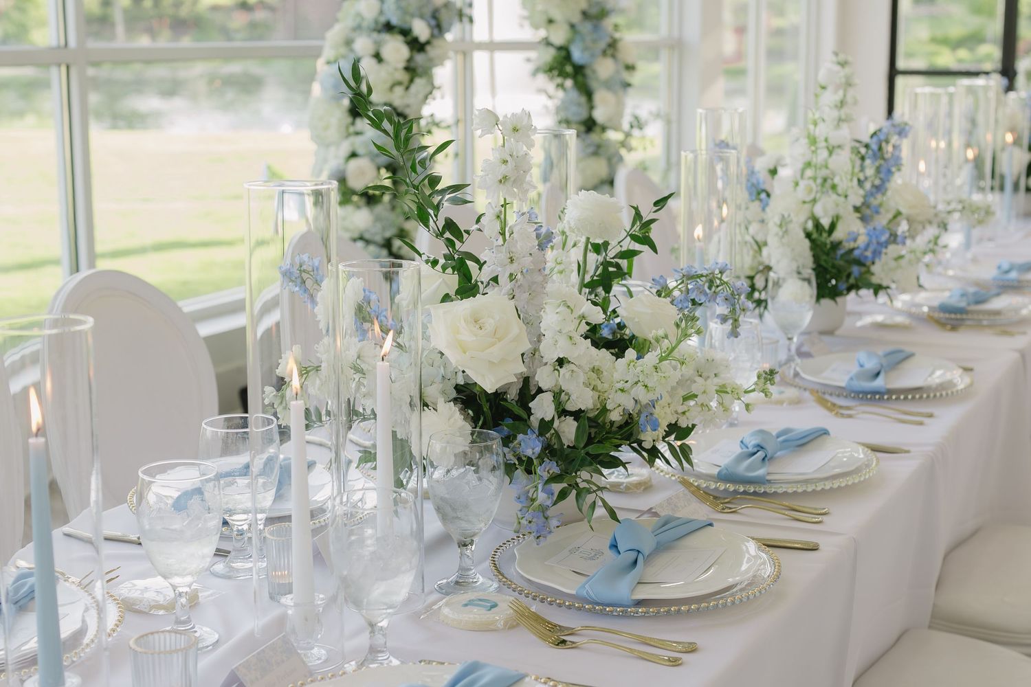 Elegant wedding reception table with white and blue floral arrangements, crystal glasses, and gold cutlery in bright venue.