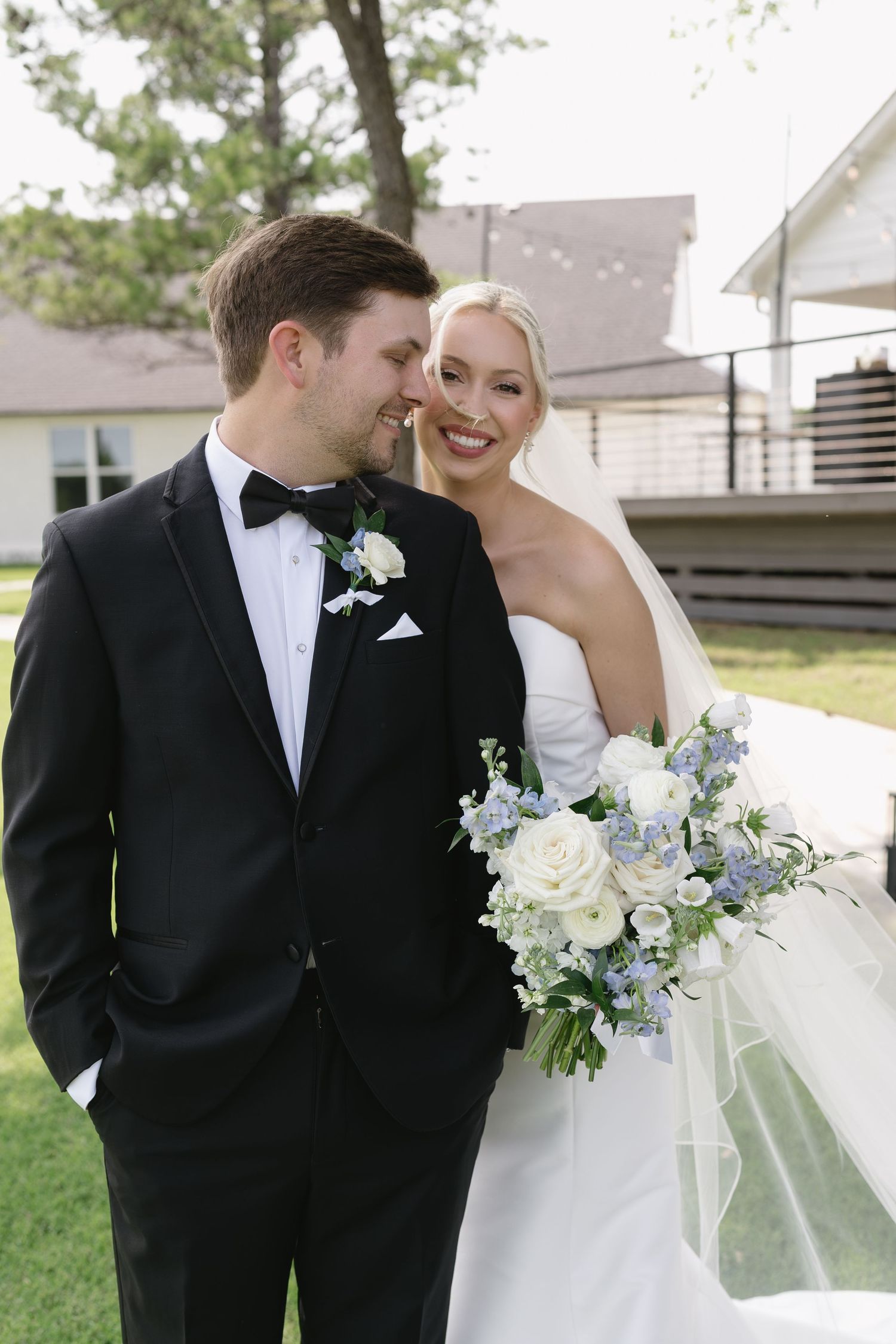 Bride in strapless gown holding blue and white bouquet smiles with groom in black tuxedo outdoors.