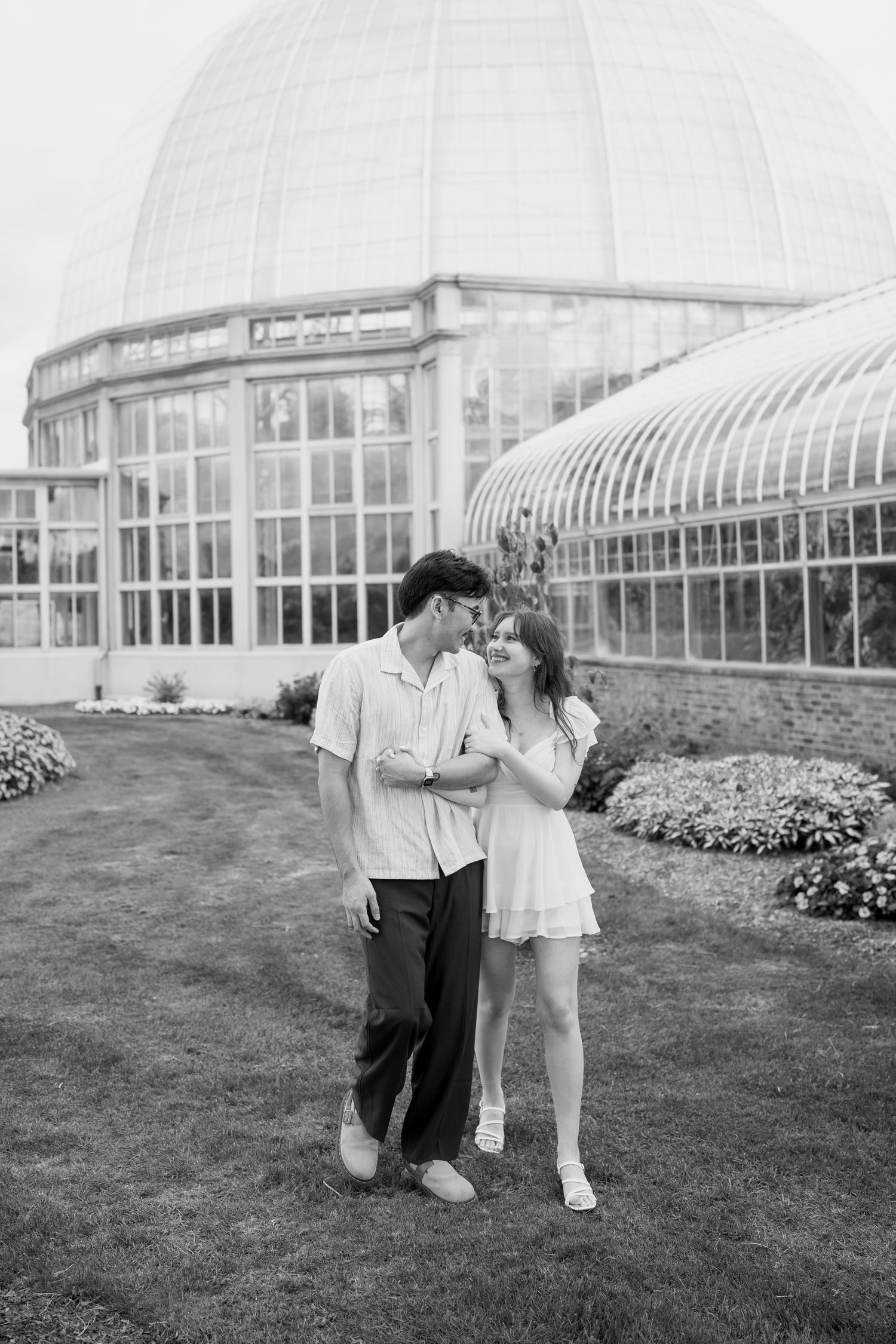 Black and white photo of a couple embracing near a glass greenhouse structure.