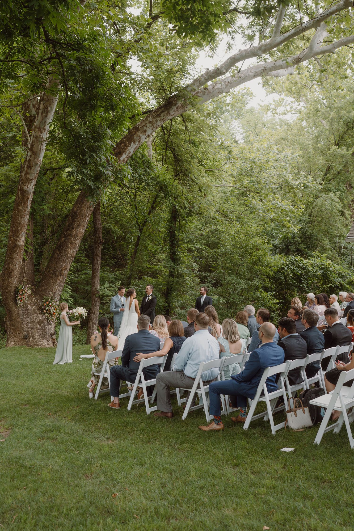 Wedding ceremony setup with white chairs arranged in rows under a canopy of green trees.