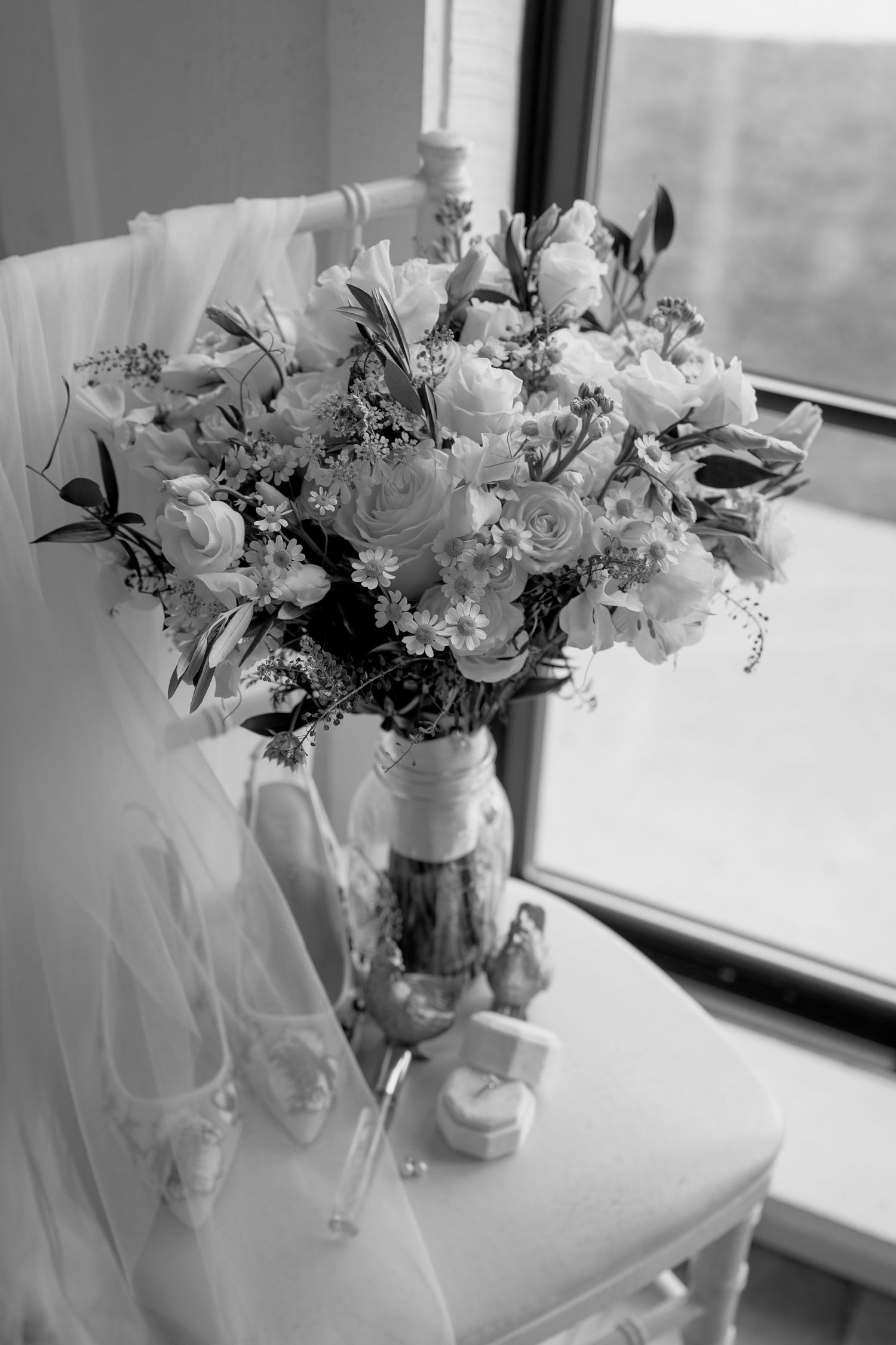 Black and white image of delicate white floral arrangements in mason jars by a window.