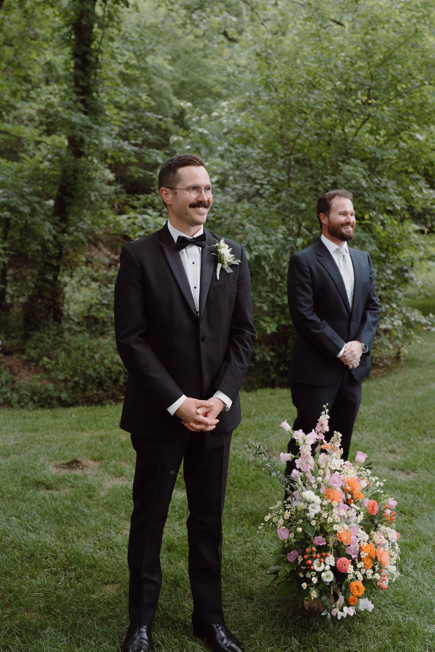 Groomsmen in black tuxedos stand together holding colorful floral bouquets outdoors.