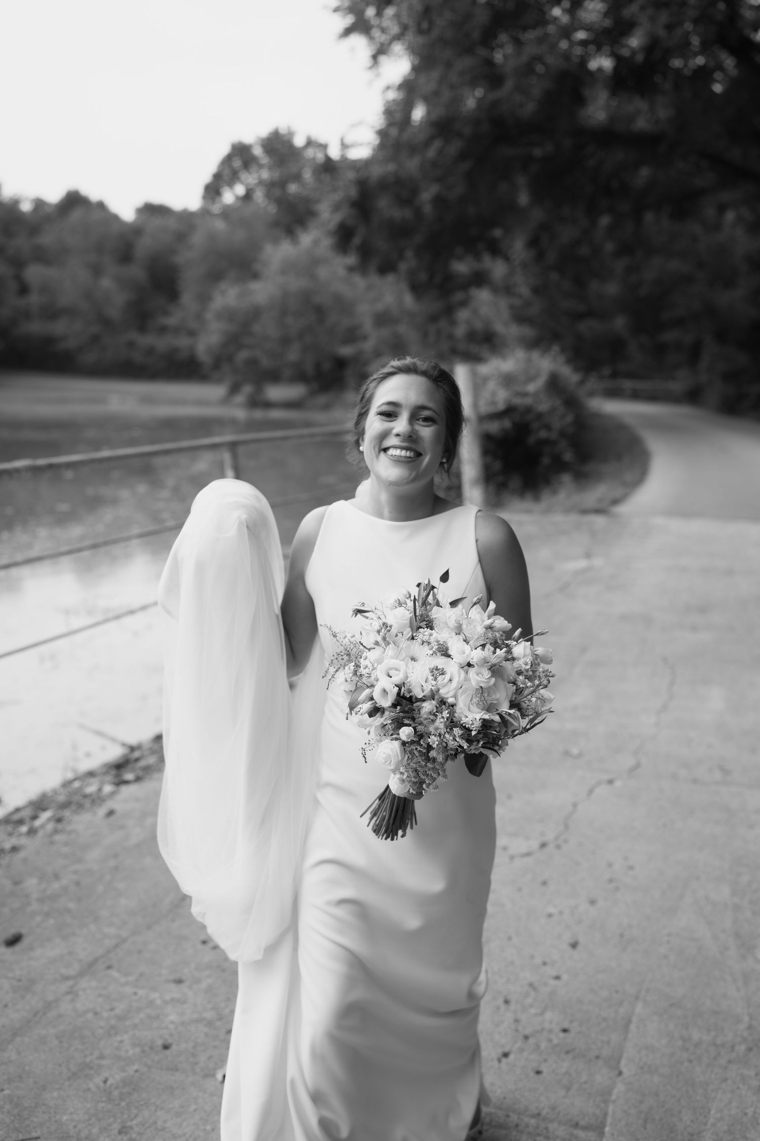 Bride in elegant white dress holds a beautiful bouquet of mixed flowers against a natural backdrop.