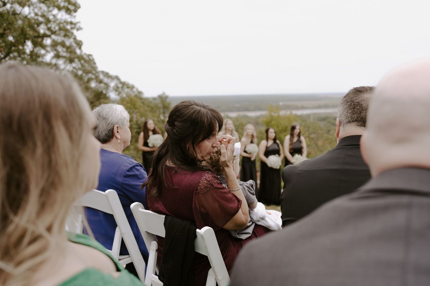 Wedding guests seated in white chairs watching outdoor ceremony from hillside vantage point.