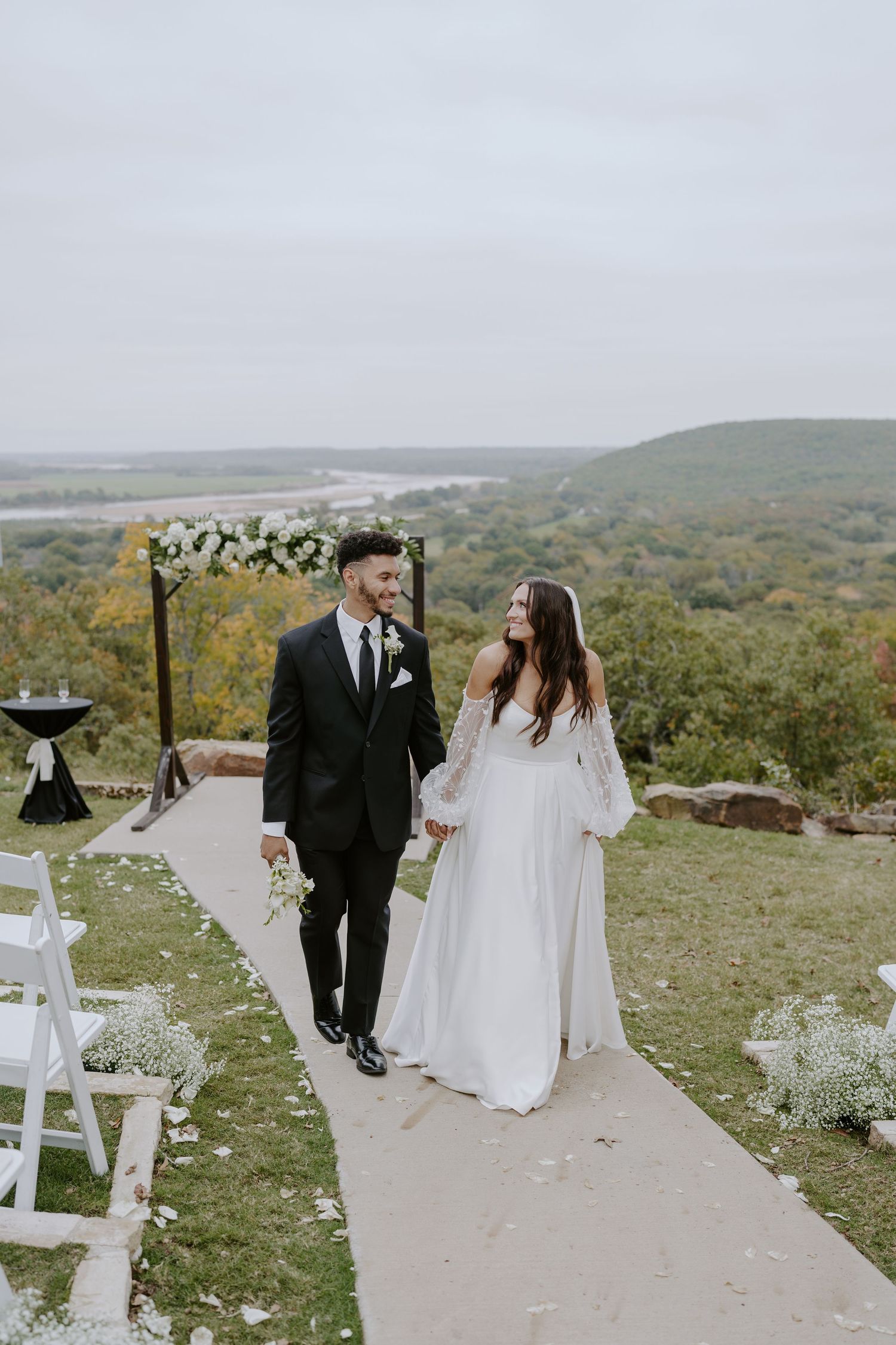 Couple walks hand in hand down an aisle lined with white chairs and floral arrangements on a hilltop.