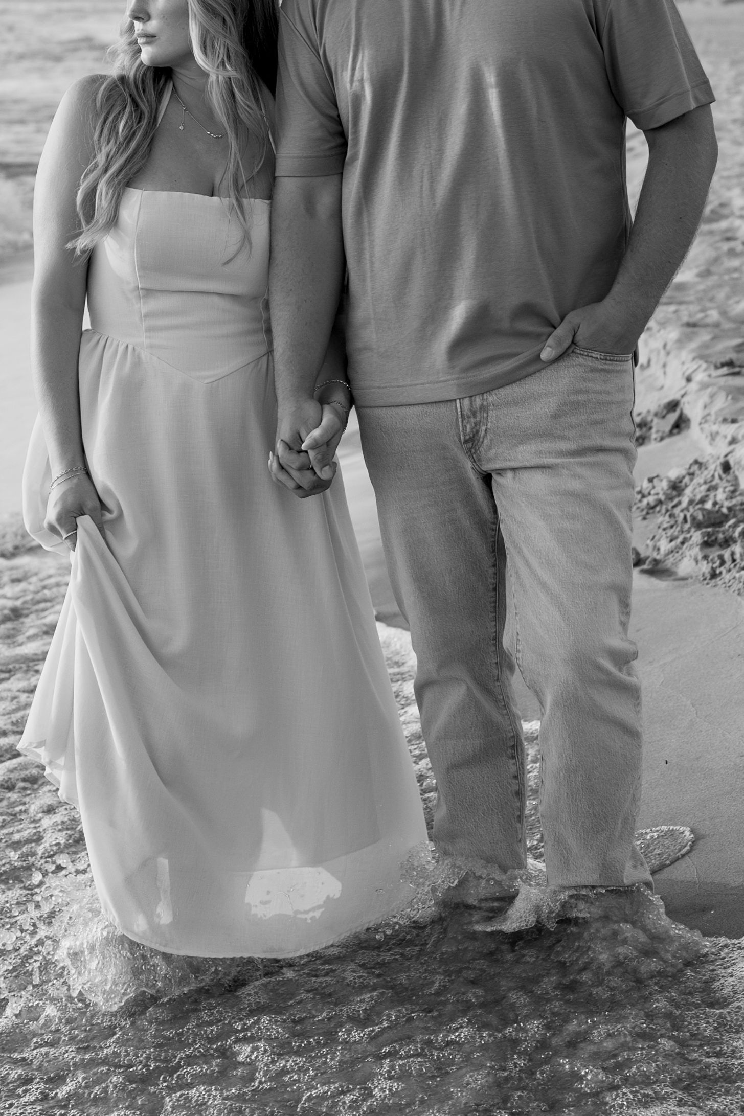 Black and white photo of a couple's casual beach stroll with waves lapping at their feet.