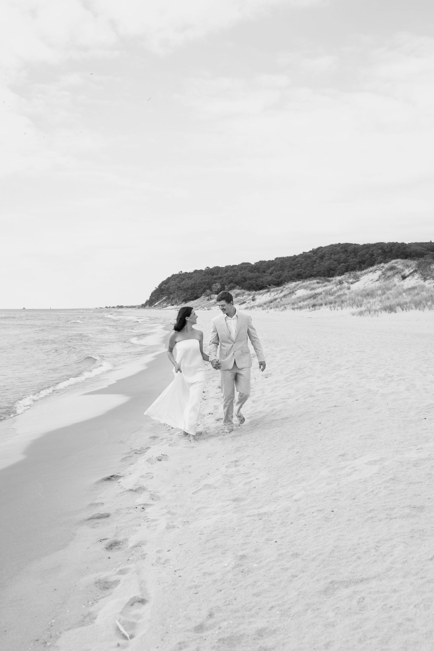Black and white photograph of a couple walking on a pristine beach with dunes in the background.