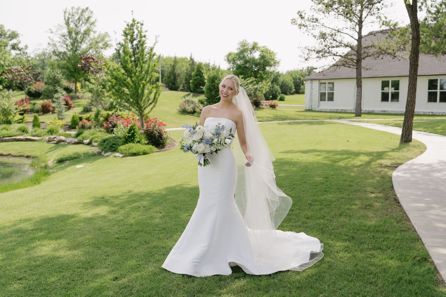 Bride in strapless mermaid gown with veil holding blue bouquet on lush green lawn near elegant venue.