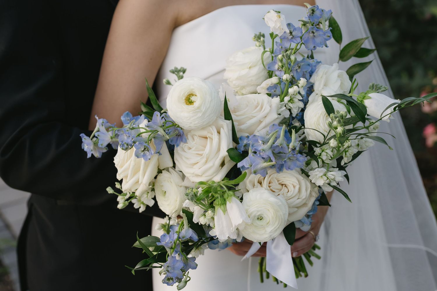Bride holding a wedding bouquet of white ranunculus and blue delphinium flowers with greenery accents.