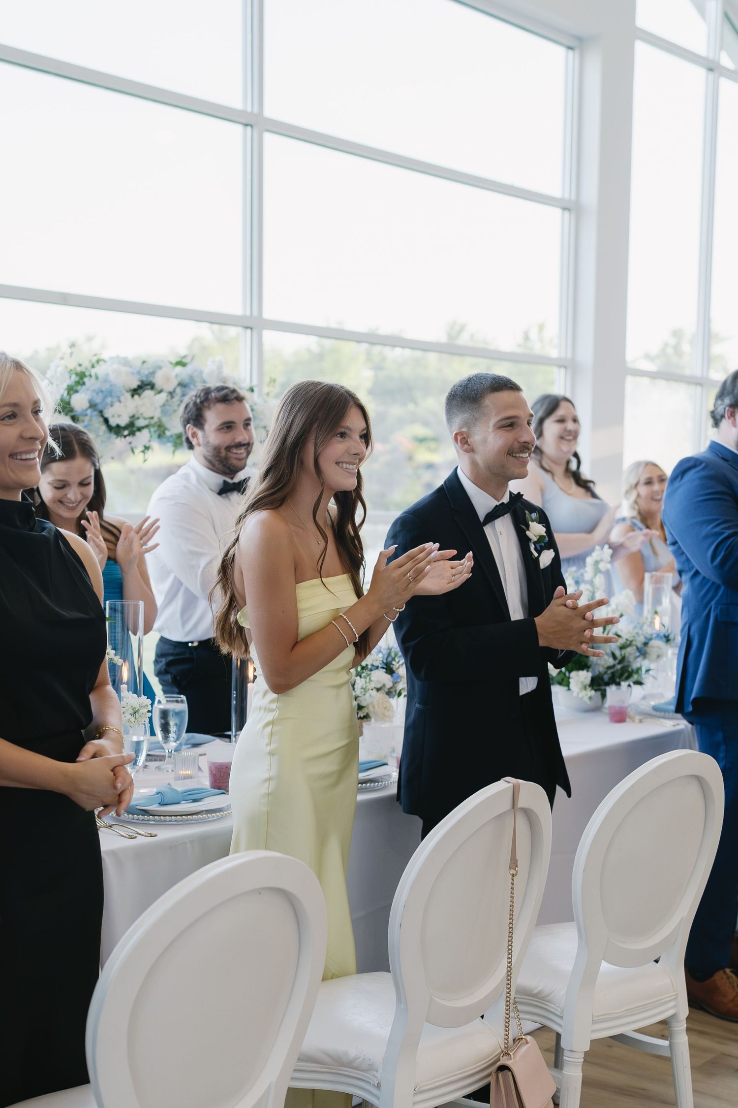 Wedding guests in formal attire clapping and smiling during a reception in an elegant venue with large windows.