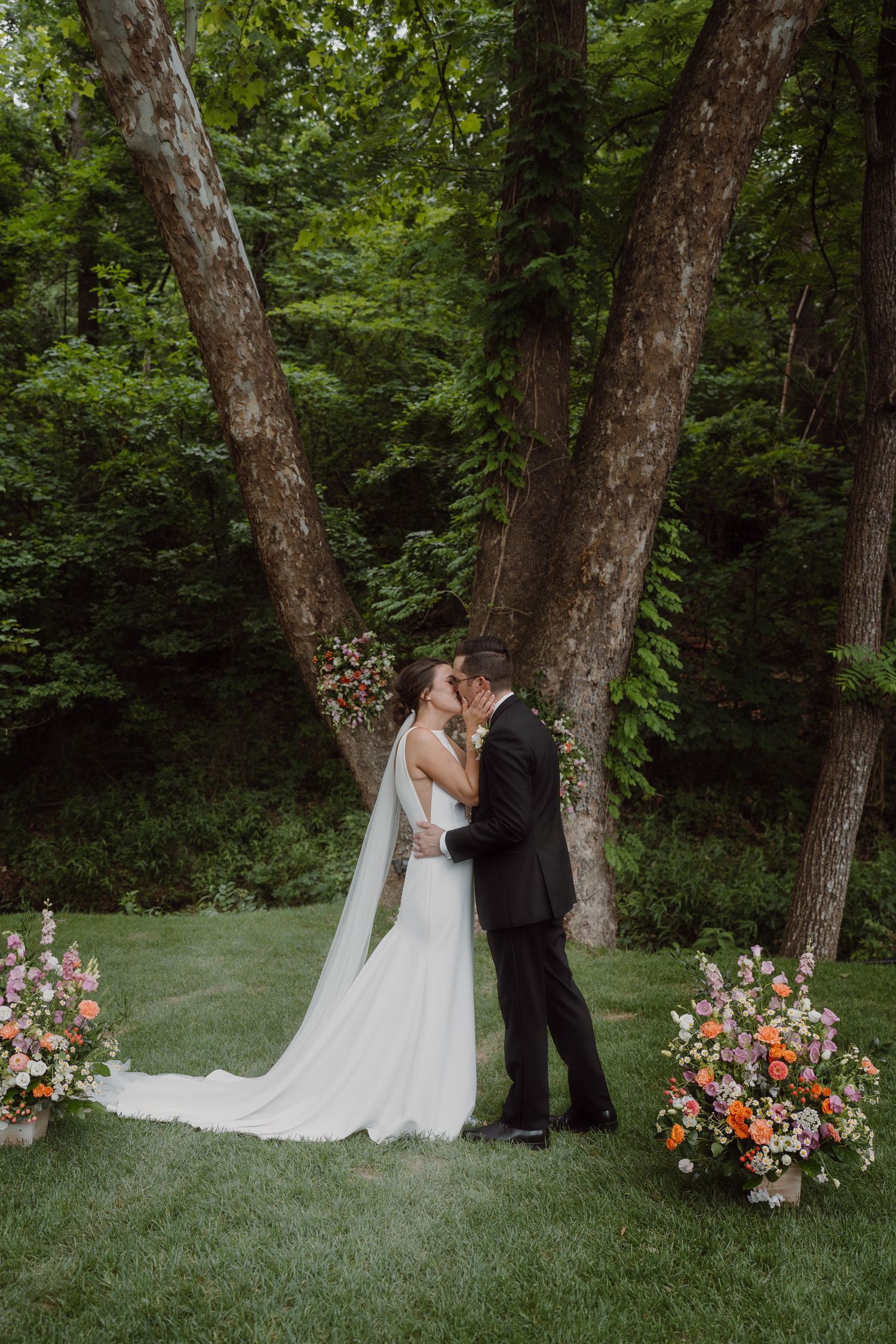 A couple shares an intimate moment between two trees decorated with colorful floral arrangements in a wooded outdoor setting.