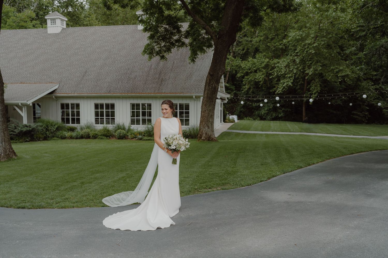 A bride in an elegant white wedding dress poses in front of a white barn building on a green lawn.